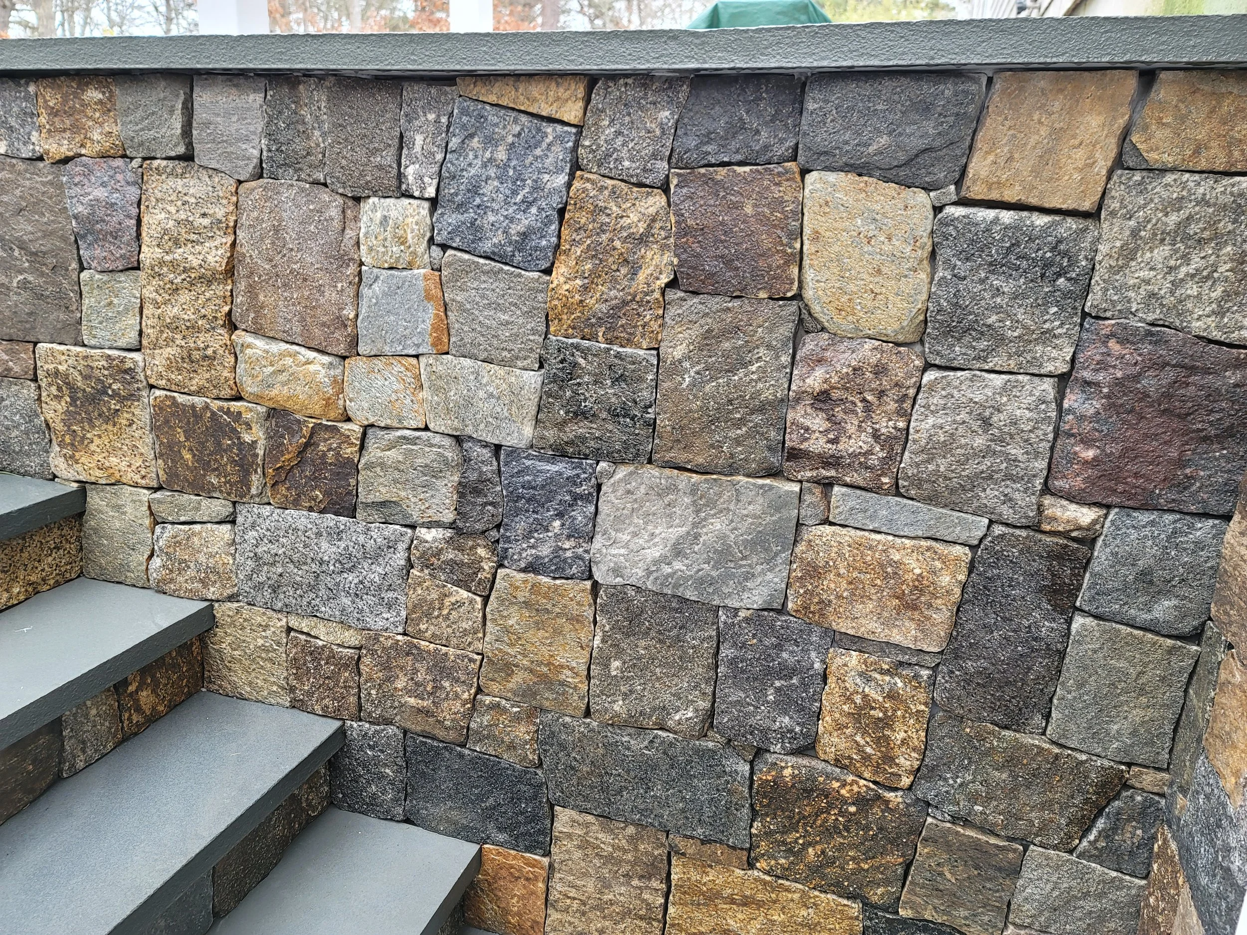 Close-up of a stone wall with irregularly shaped stones in various shades of brown, gray, and black, and steps made of smooth gray concrete at the bottom left.