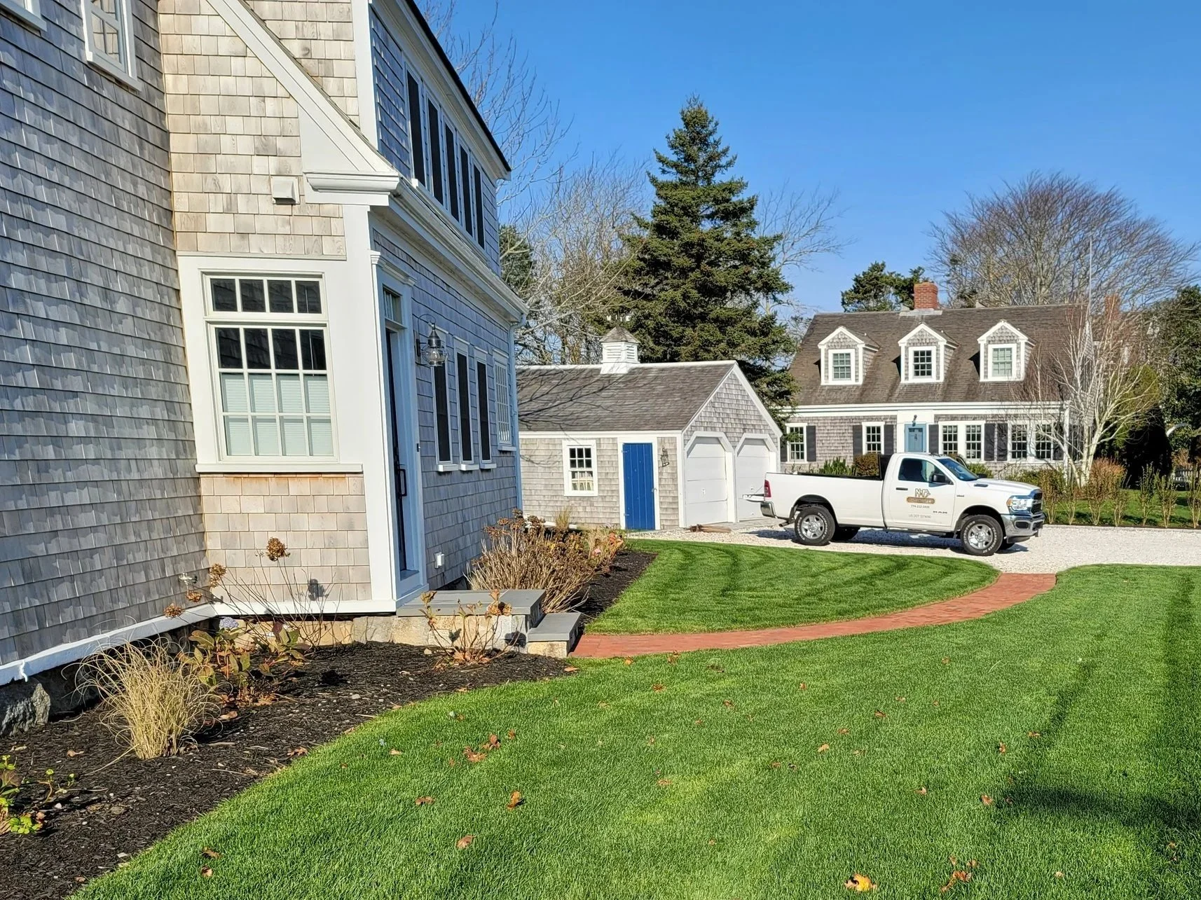 Front yard of a house with well-maintained green grass, blooming garden beds, a gravel driveway, a white pickup truck parked, and neighboring houses with similar architecture in the background under a bright blue sky.