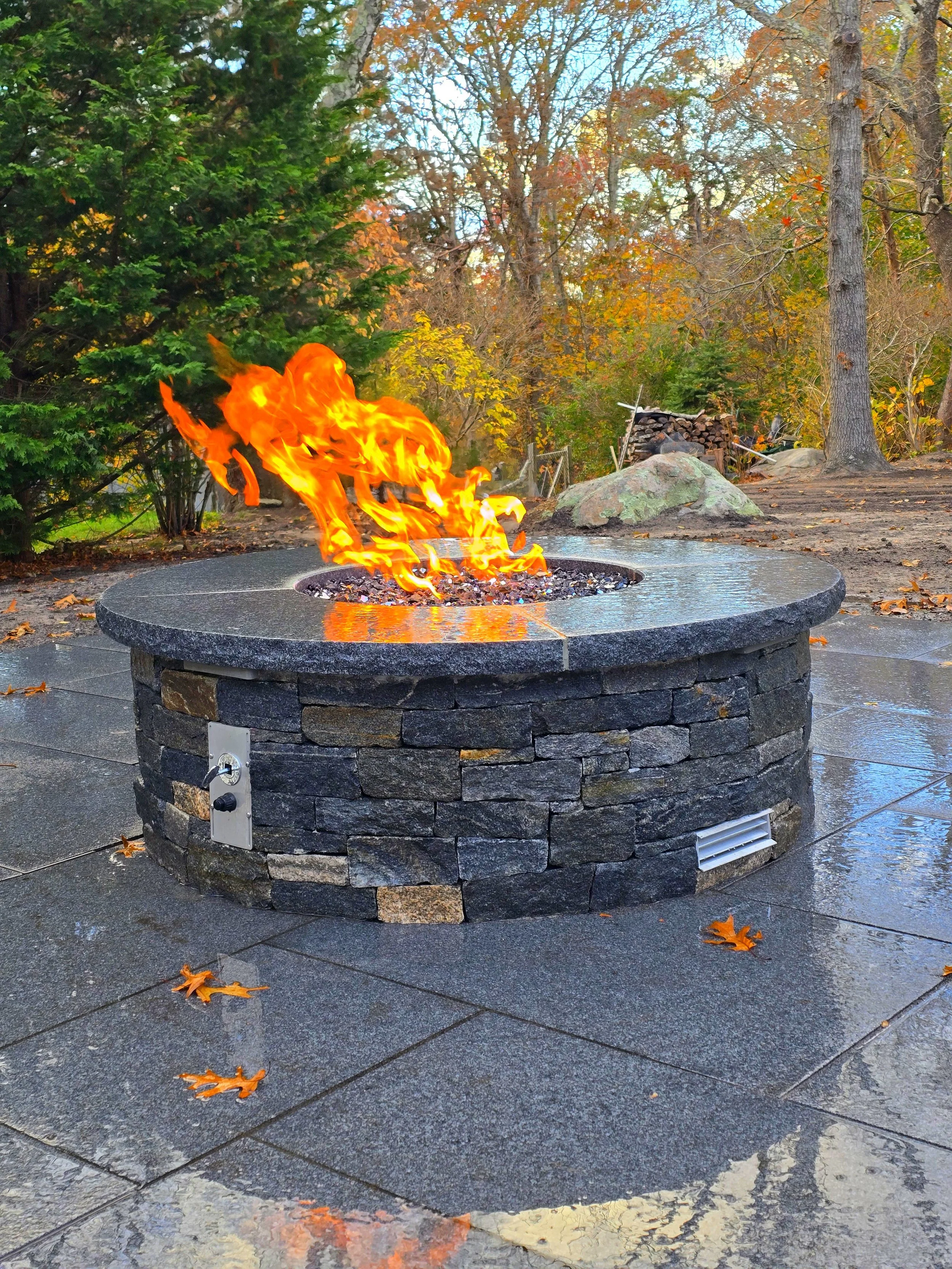 Outdoor fire pit with flames, surrounded by wet stone tiles, trees with autumn foliage, and a stack of firewood in the background.