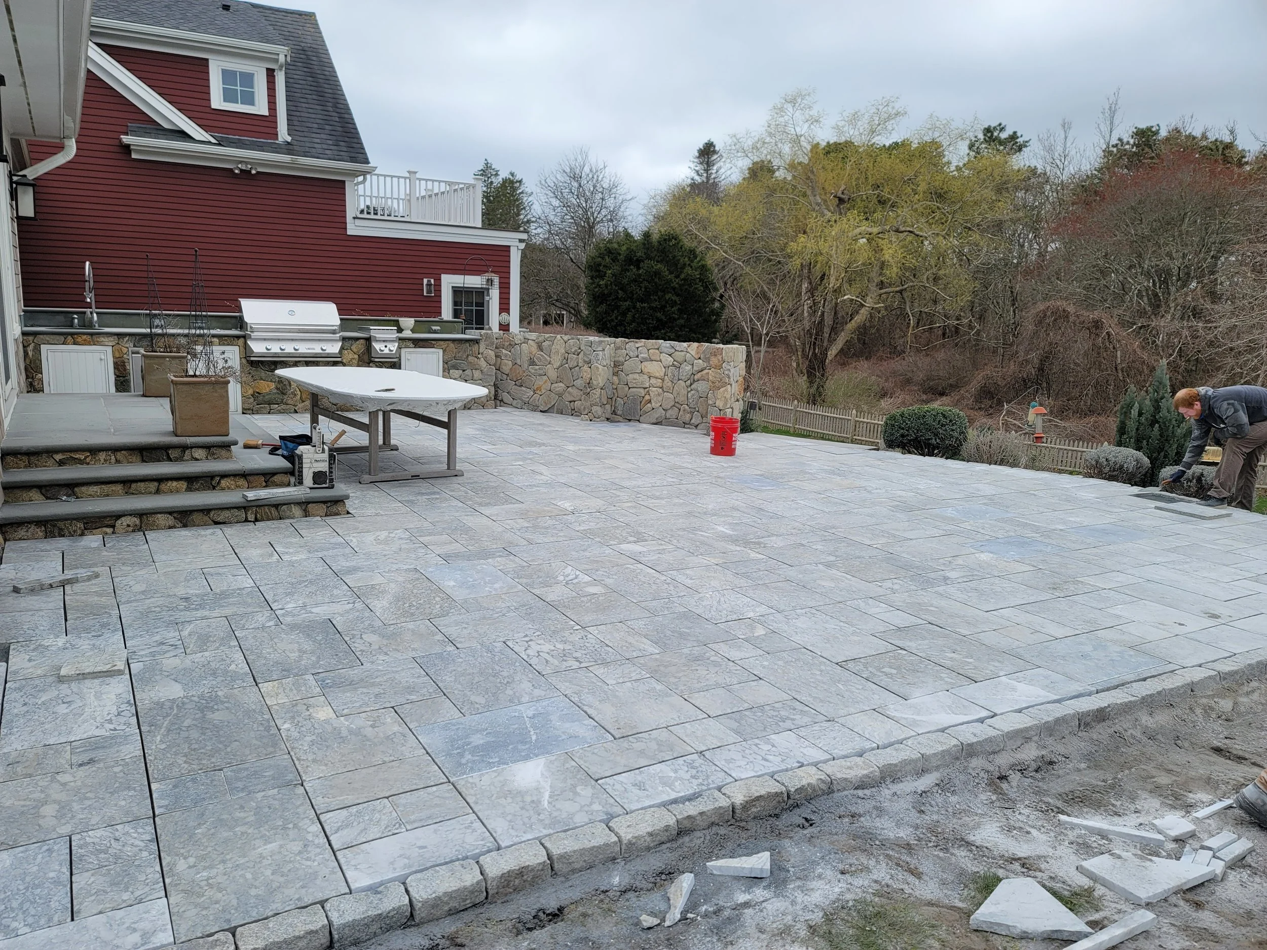 A person installing stone pavers on a backyard patio, with outdoor furniture and a barbecue grill, surrounded by a stone retaining wall and trees in the background.