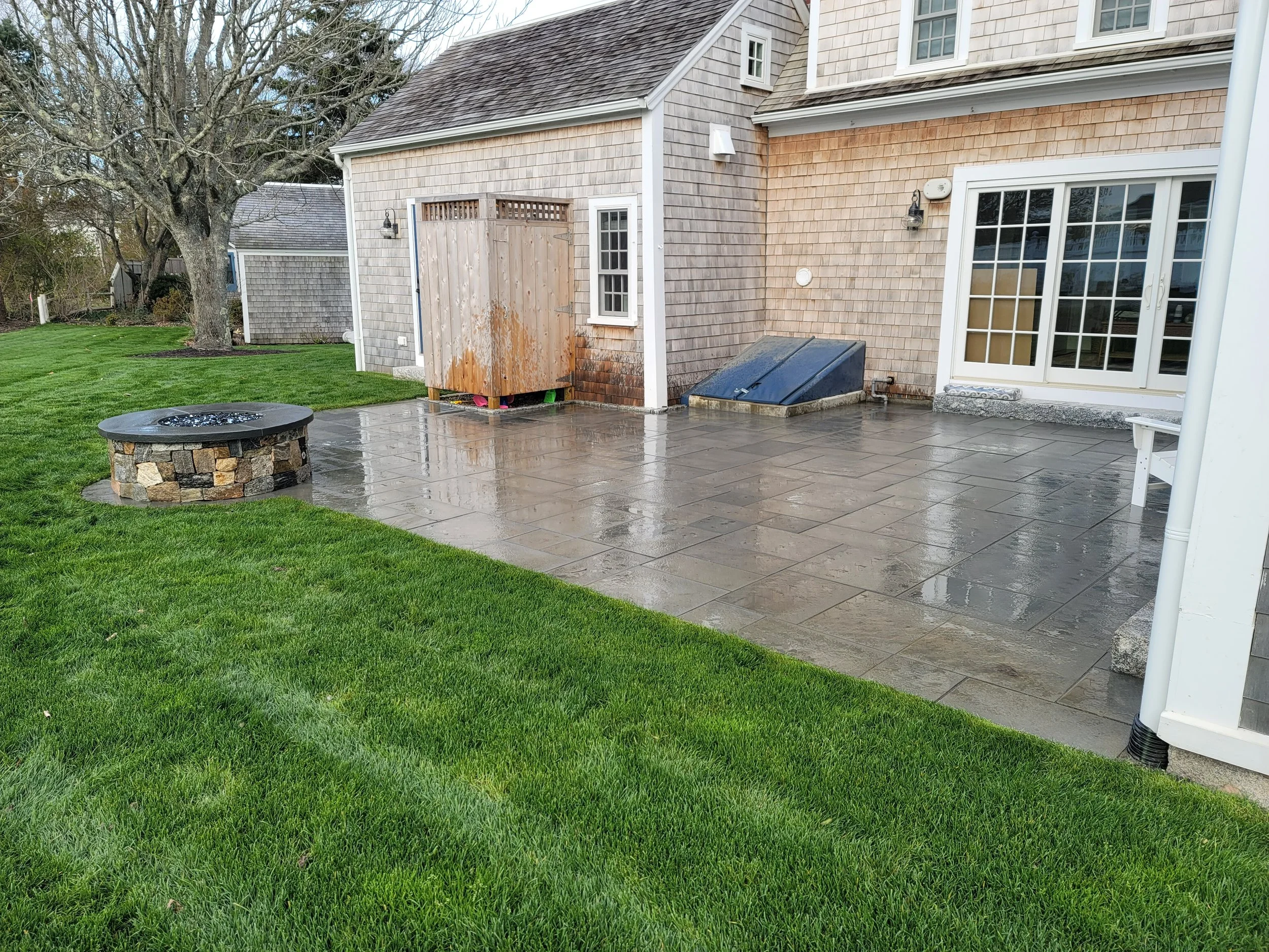 A backyard patio area with wet concrete paving stones, a fire pit with a stone base, a small wooden shed with rusted bottom, a black box, a white sliding door, and green grass, with a tree and neighboring house in the background.