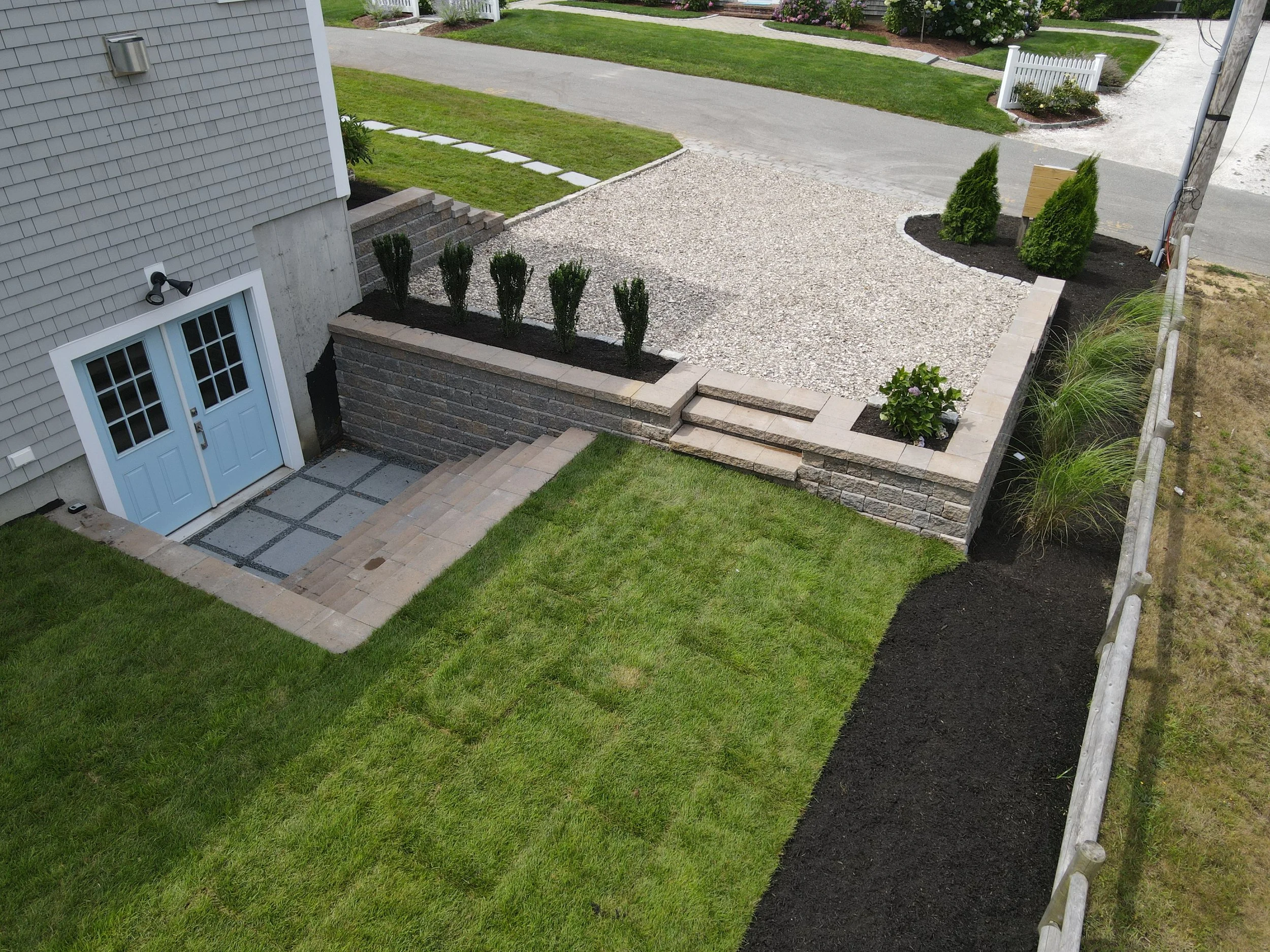 An aerial view of a landscaped backyard with a small green lawn, stone steps, a raised garden bed with shrubs, and a gravel area with a bench and additional plants, bordered by a fence and sidewalk.