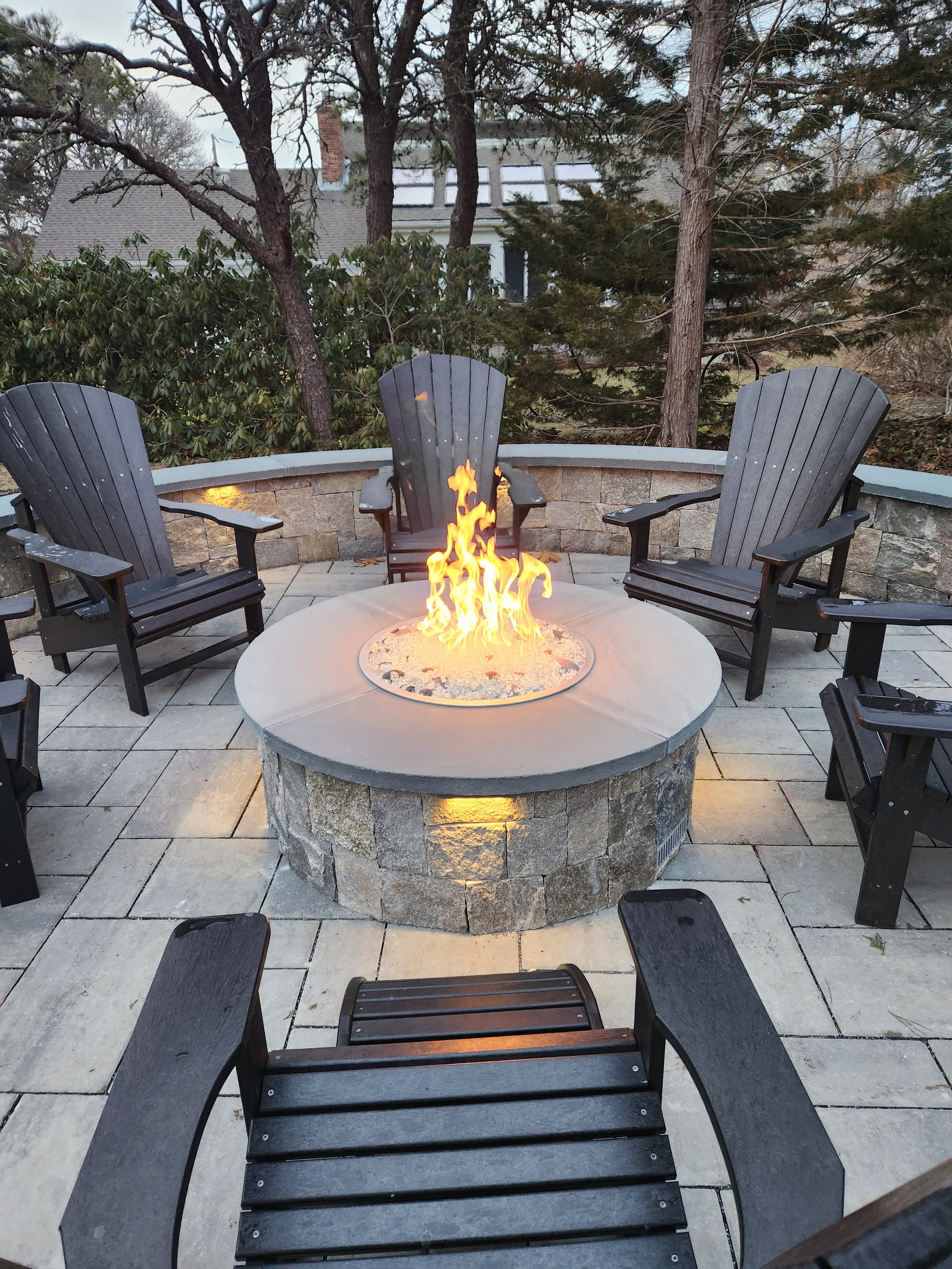 Outdoor patio with a fire pit surrounded by black Adirondack chairs and a curved stone wall, with trees and house in the background.