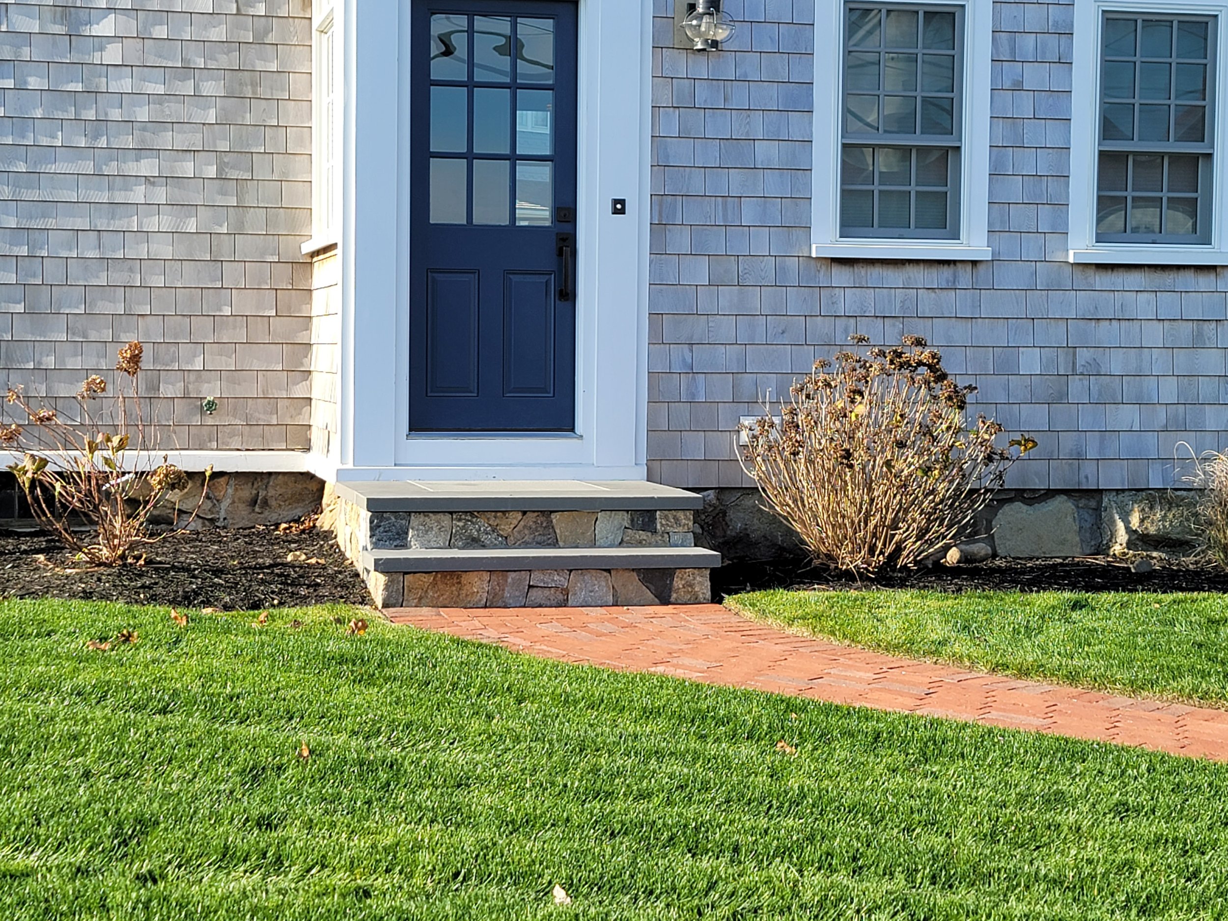 Front door entrance with steps, brick pathway, neatly trimmed grass, and bushes on the side of a house with shingle siding and double-hung windows.