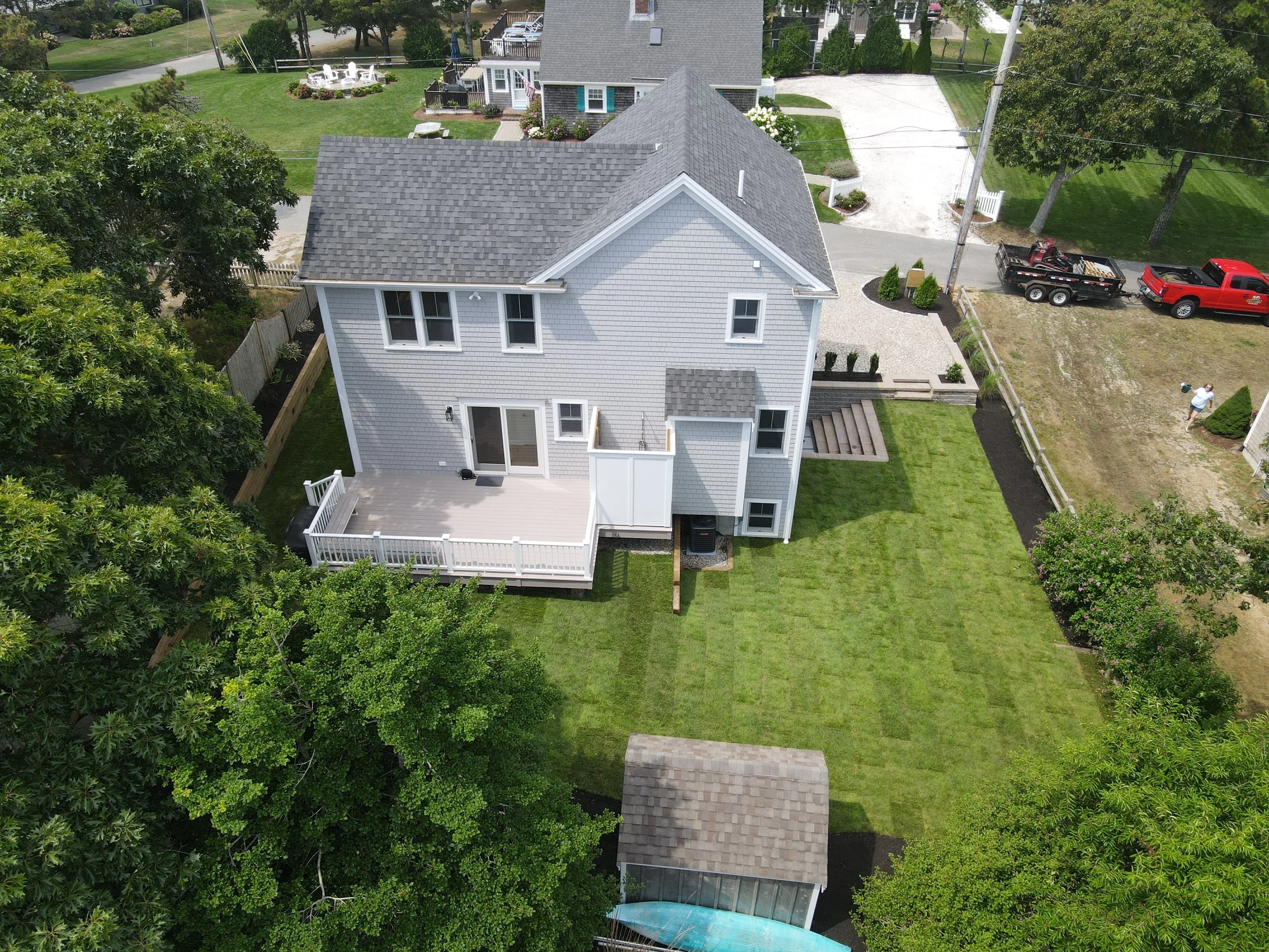 Aerial view of a two-story house with a gray roof and white siding, a backyard with a deck, grassy lawn, and trees, neighboring houses, a driveway with a red truck and trailer, and a person walking nearby.