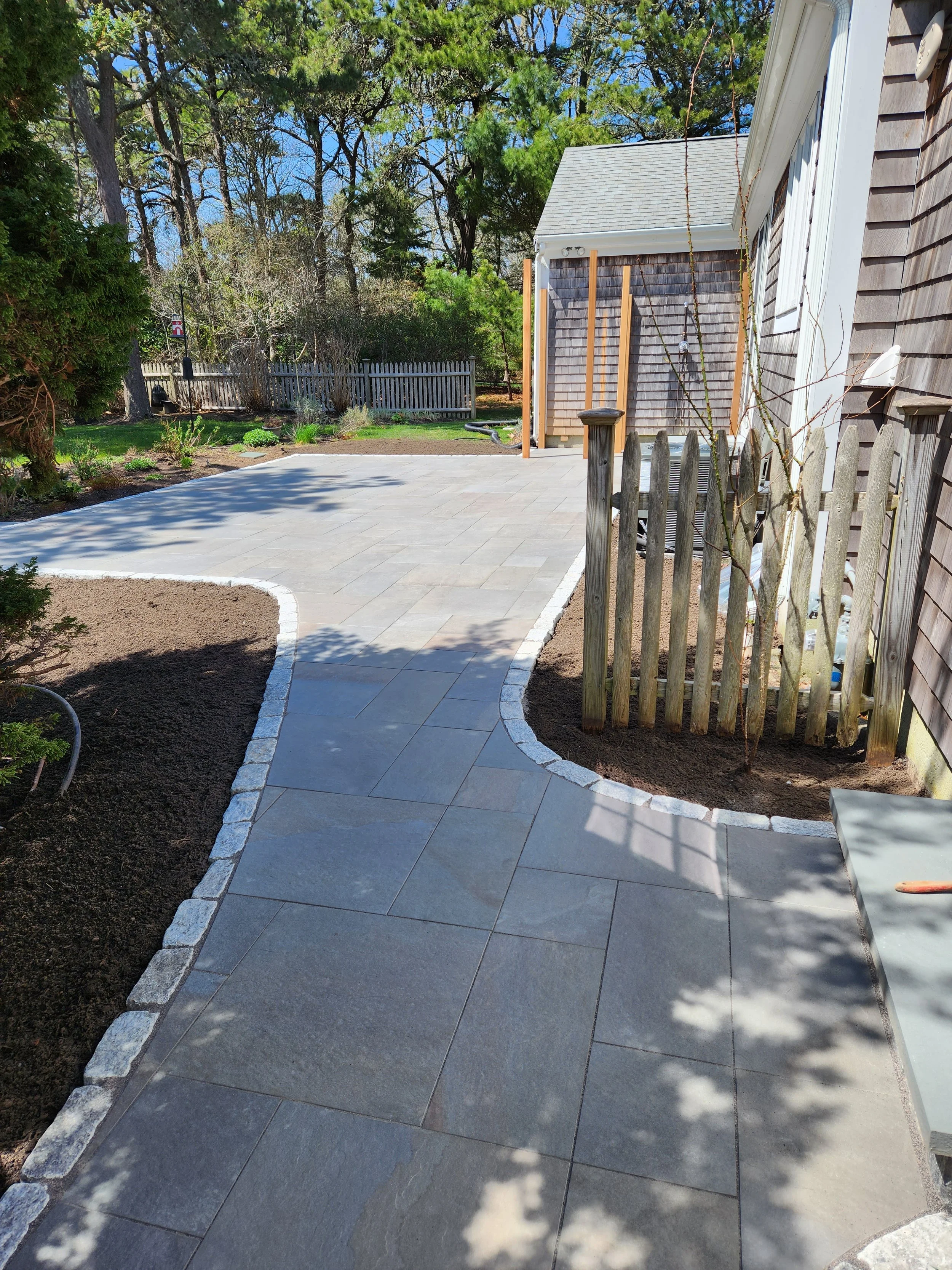 A newly paved backyard patio with gray stone tiles, bordered by white stones. There is a wooden fence, some trees with shadows, and part of a house with siding visible.