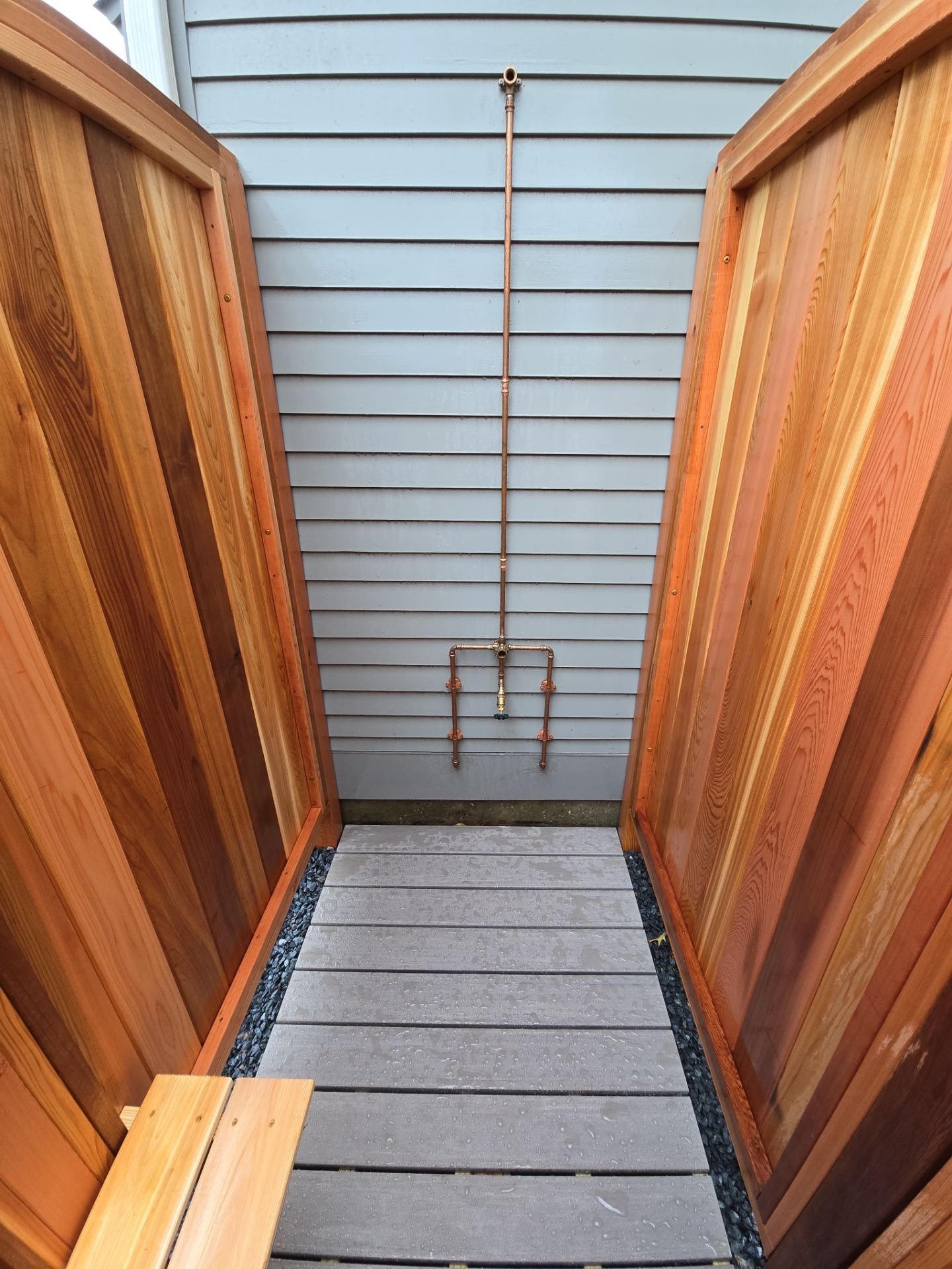 Small outdoor shower area with wooden fencing, gray flooring, and exposed plumbing pipes on a blue wall.