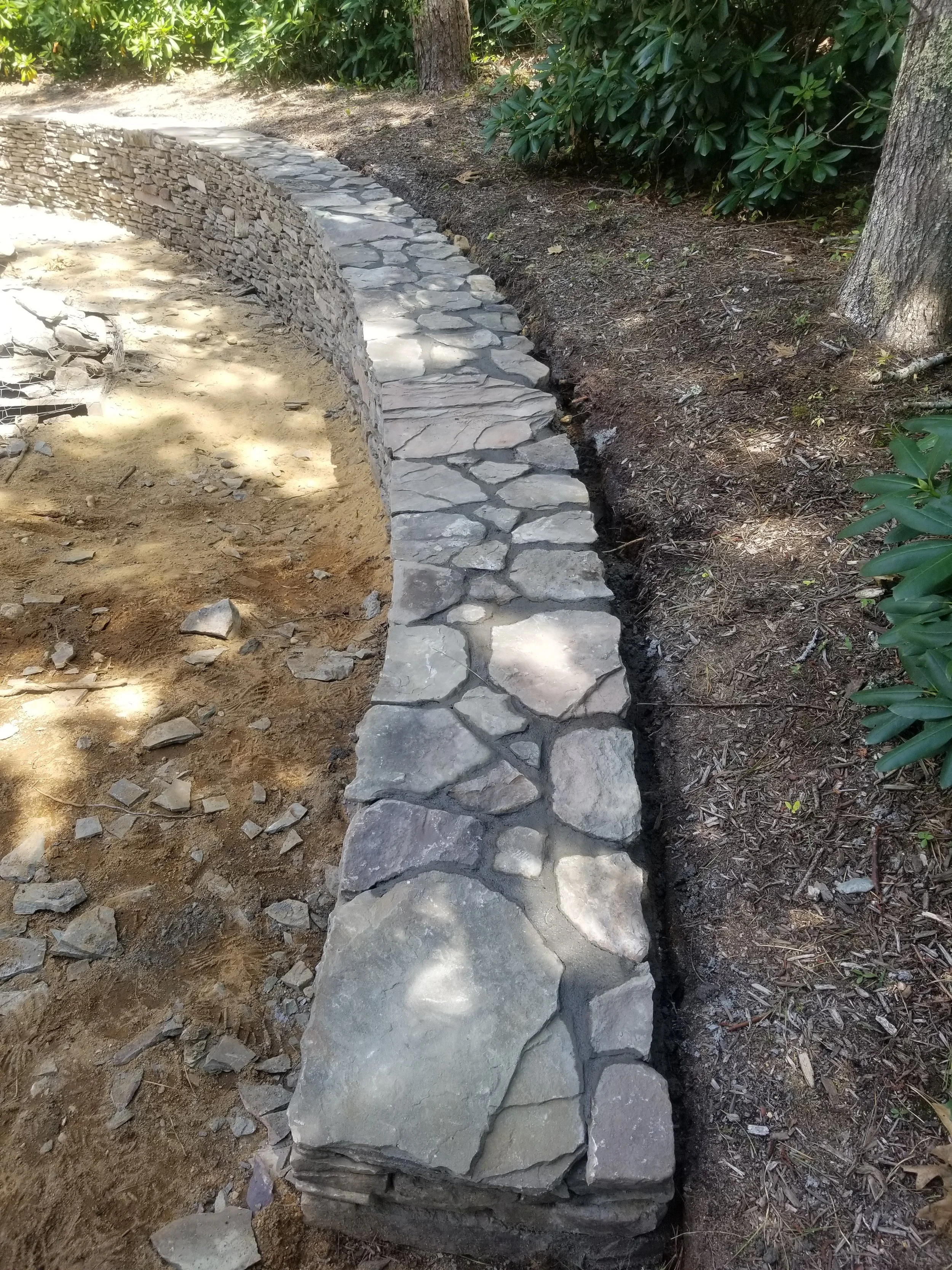 A curved stone walkway under construction, with large flat stones laid along the edge and smaller stones filling the interior, bordered by dirt and green shrubs.