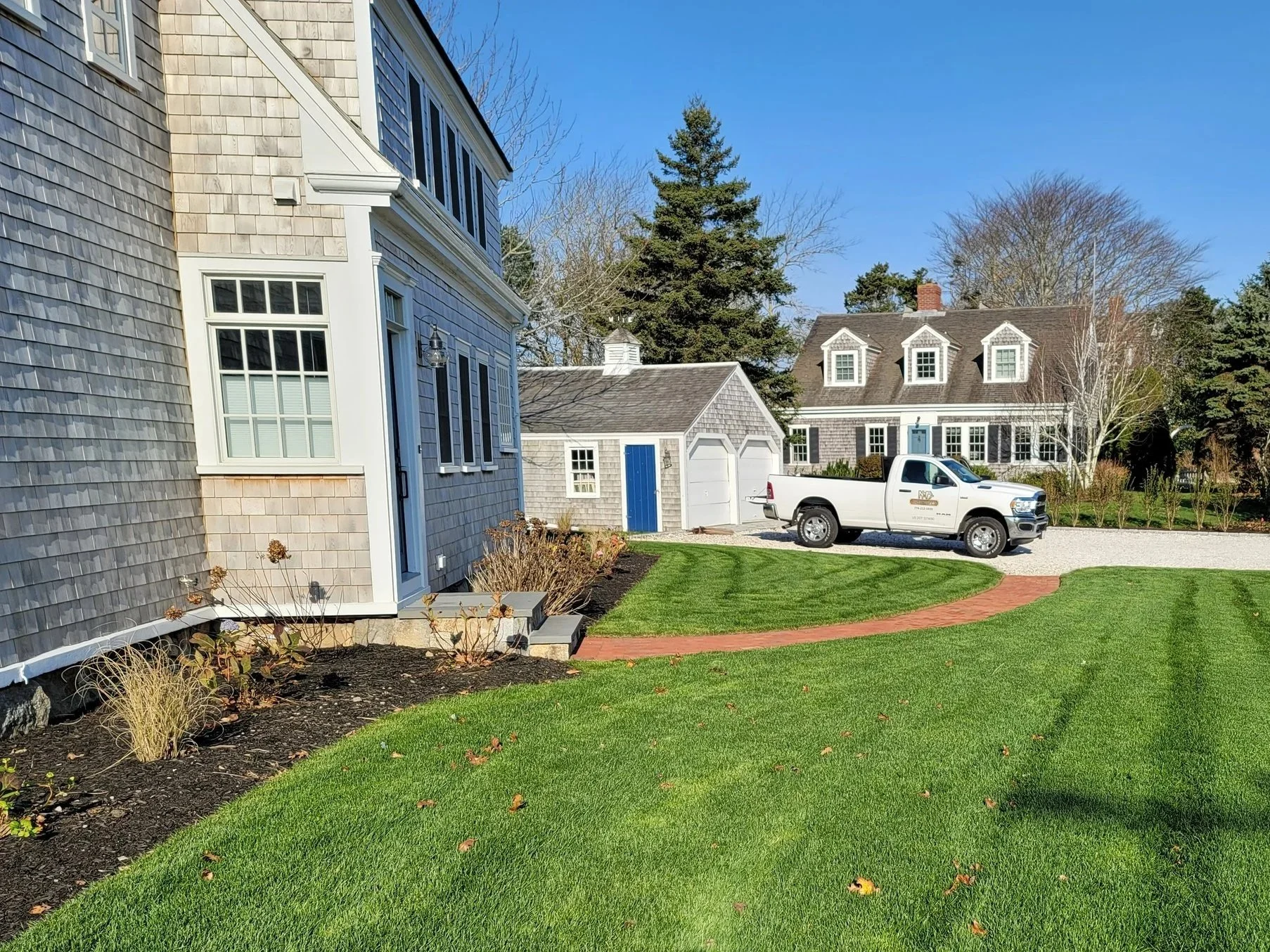 A yard with green grass, a brick pathway, and two houses in the background, one with gray siding and a smaller shed with a white door. A white pickup truck is parked on a gravel driveway.