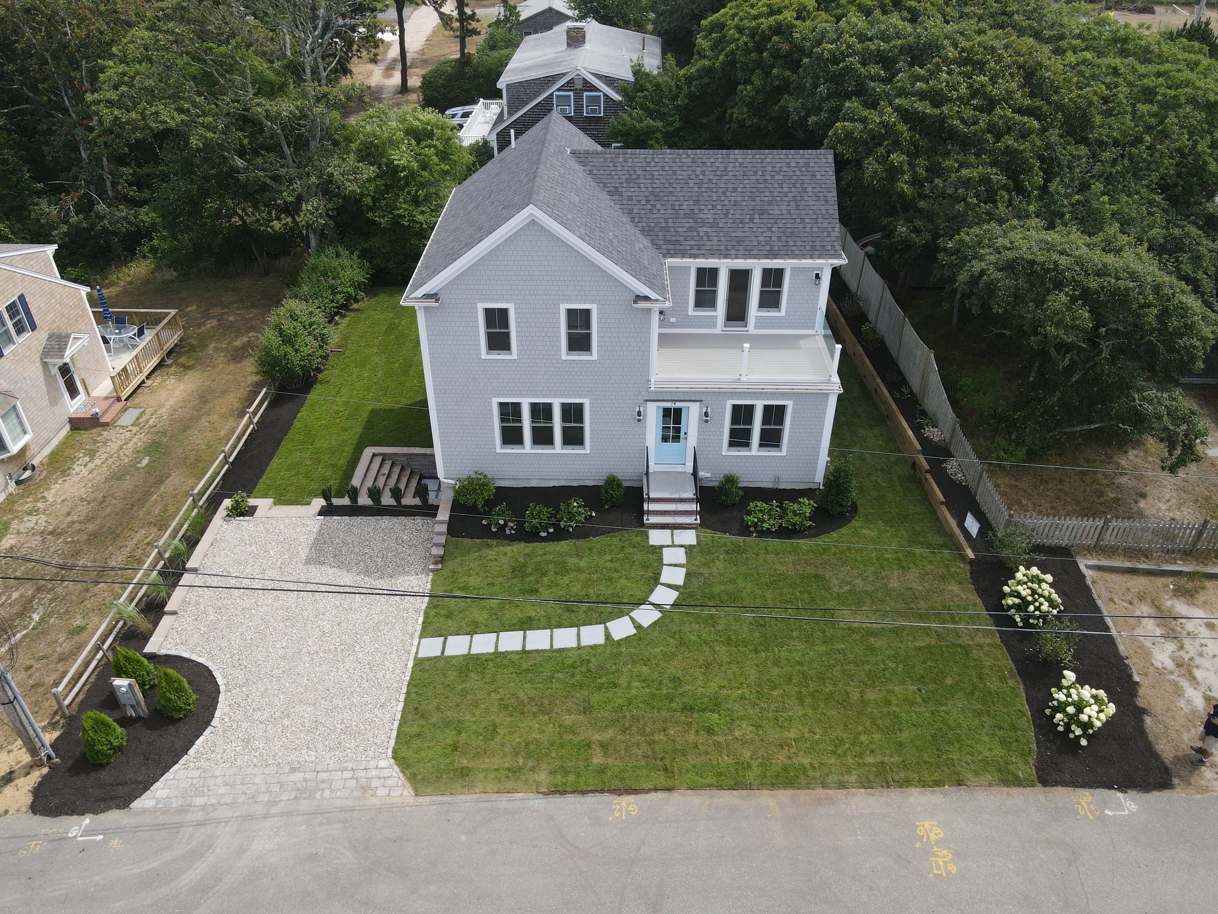 Aerial view of a two-story light gray house with a front porch, surrounded by a neatly landscaped lawn with a curved pathway and flower beds, fenced backyard, and neighboring houses with trees in the background.