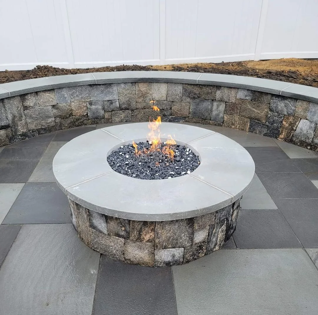 Outdoor stone fire pit with small flames in the center, surrounded by a gray stone patio and a curved retaining wall of stacked stones, with a white fence in the background.