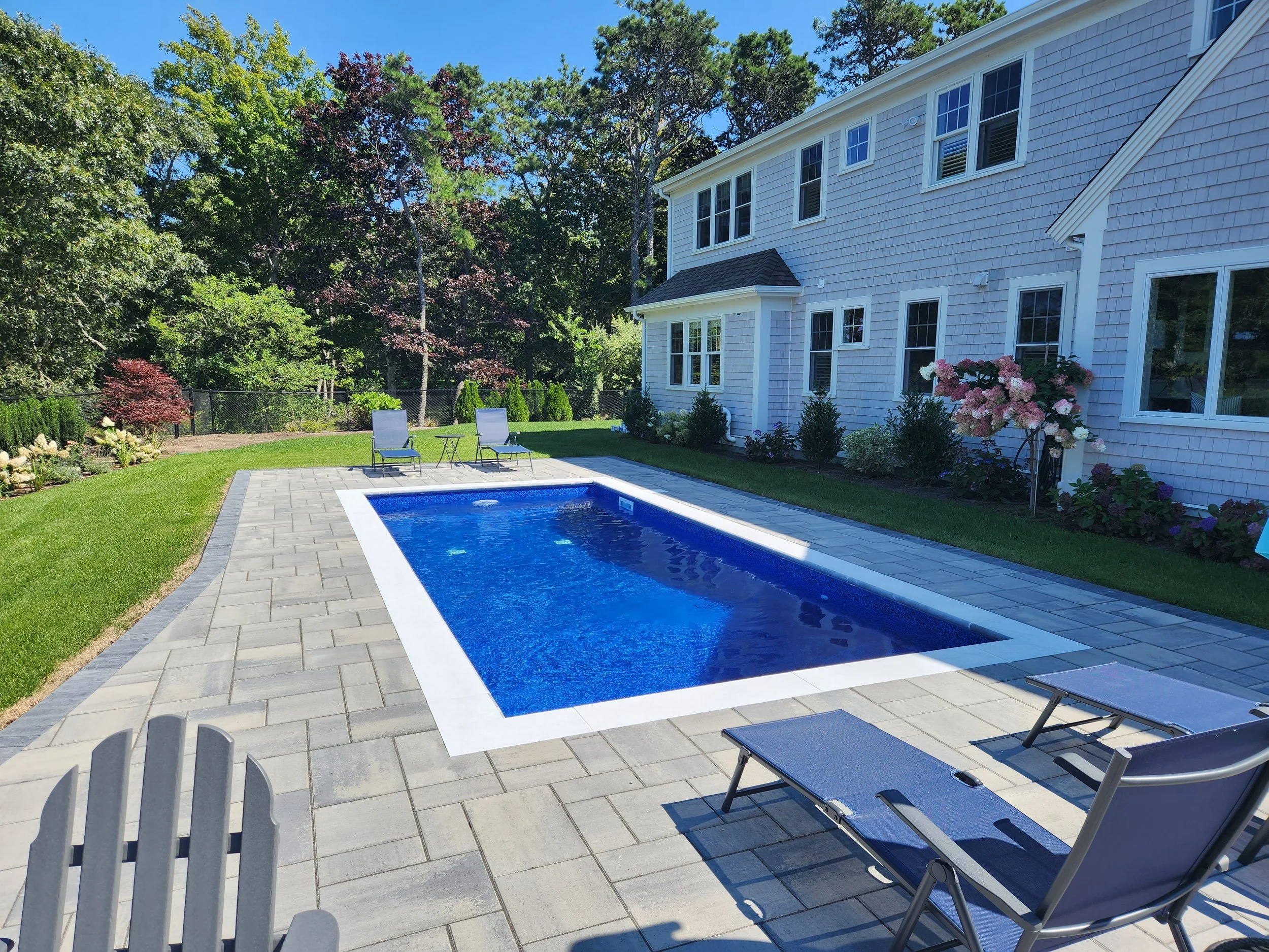 Backyard with in-ground swimming pool surrounded by gray stone patio, lawn, and garden beds with flowers and shrubs, next to a white house with multiple windows.