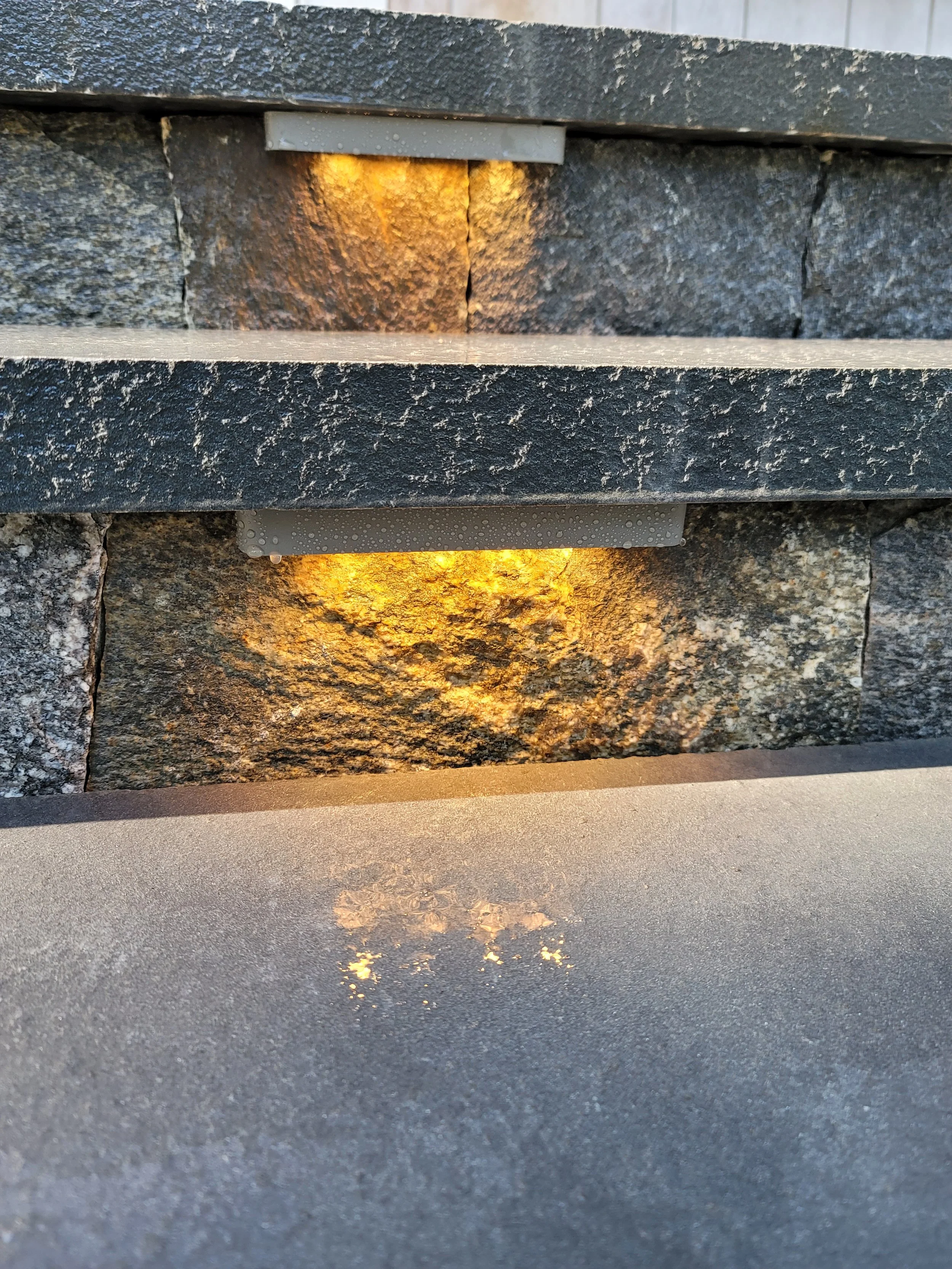 Close-up of a stone wall with a small, rectangular outdoor light illuminating the stone, and its reflection on a flat surface below.