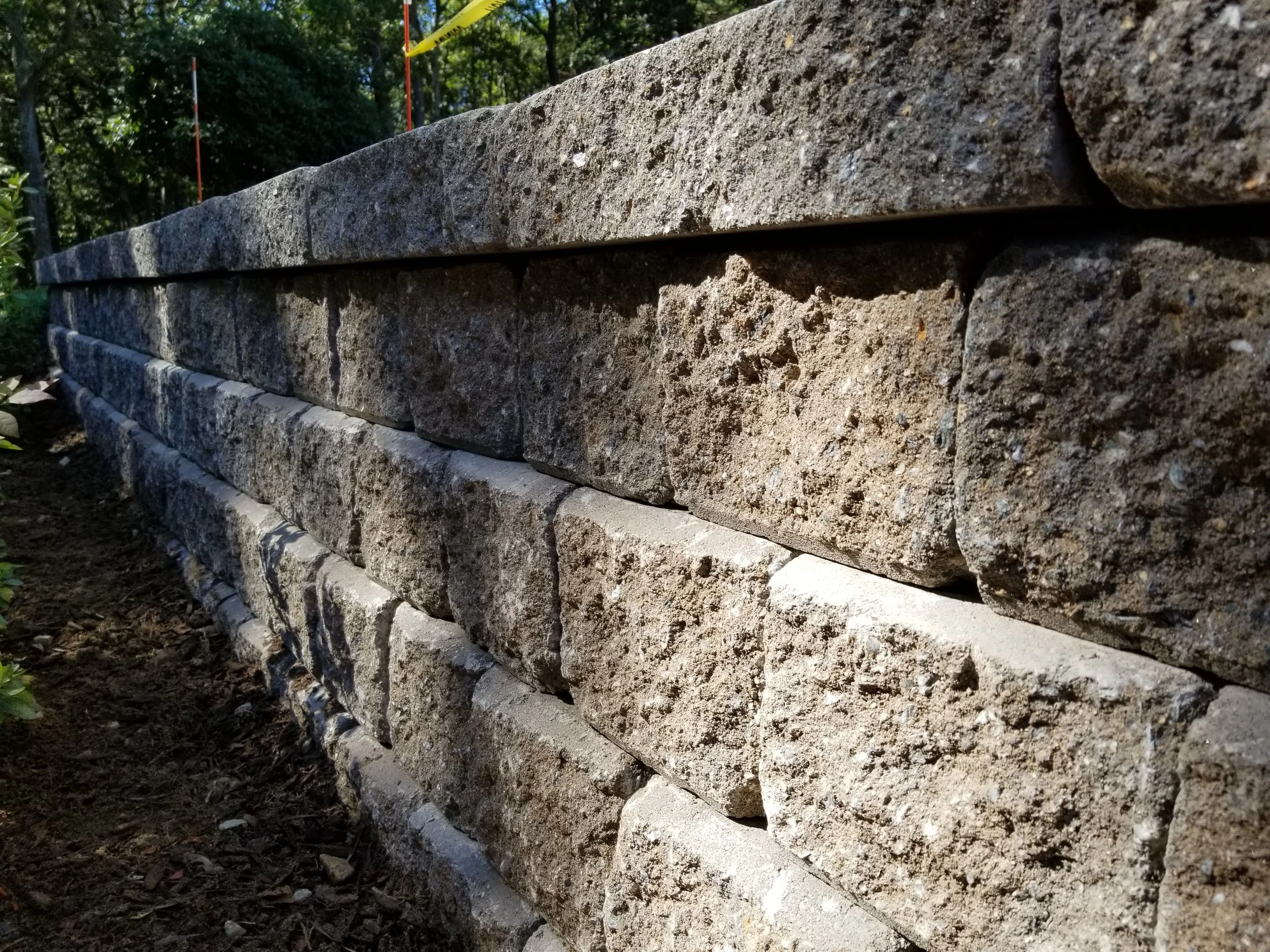 Close-up of a layered stone retaining wall with landscaping soil and trees in the background.