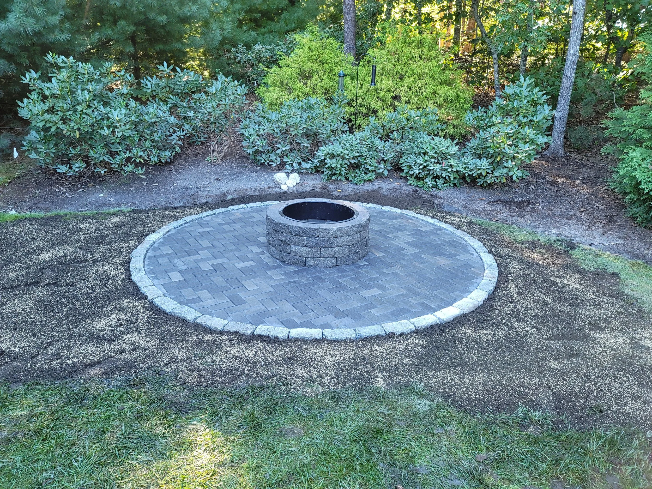 A circular brick fire pit on a paved area surrounded by a garden with bushes and trees.