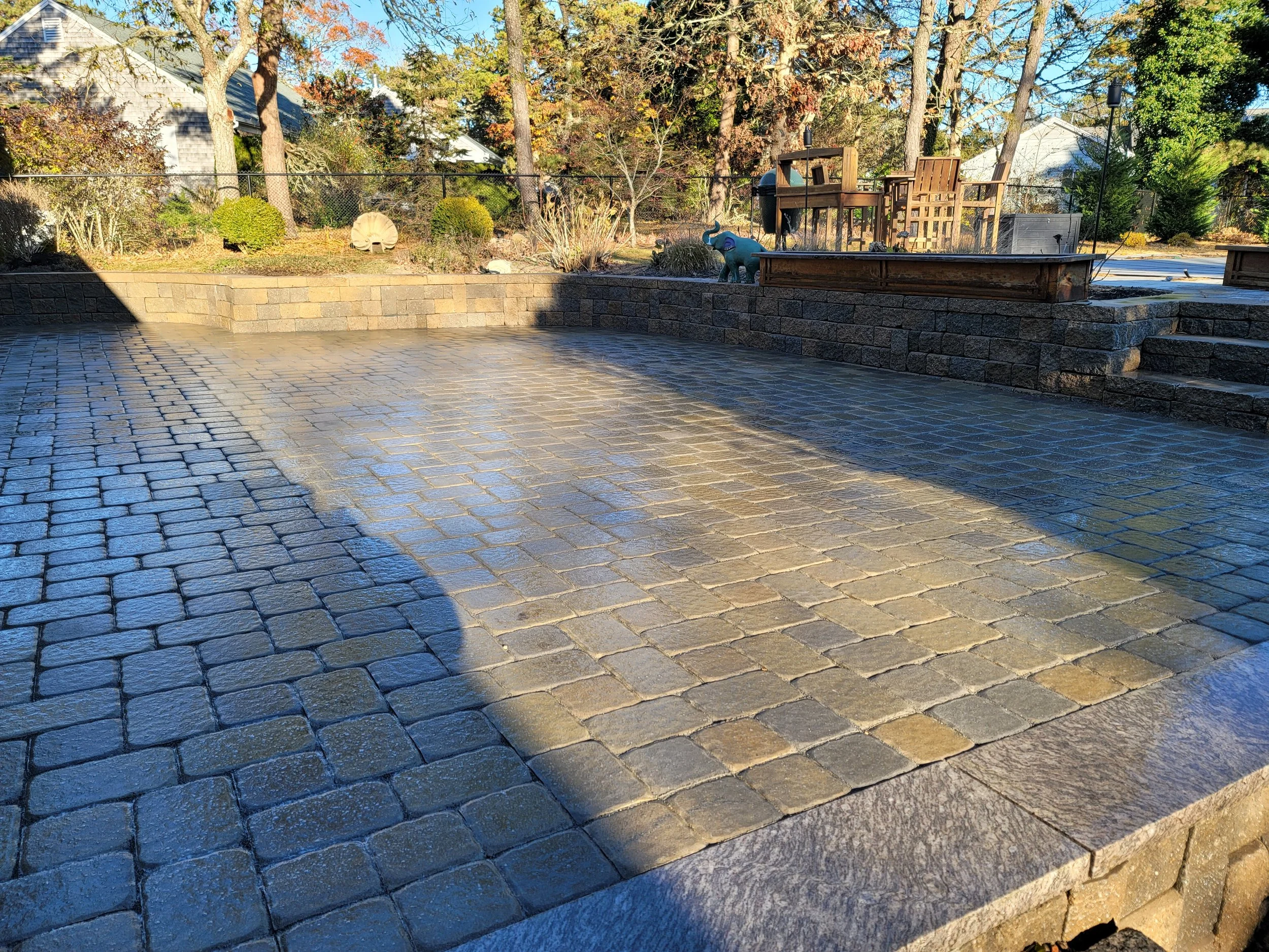 Patio with cobblestone pavers in sunlight and shadow, backyard scene with trees, bushes, wooden furniture, a dog statue, and a stone planter.
