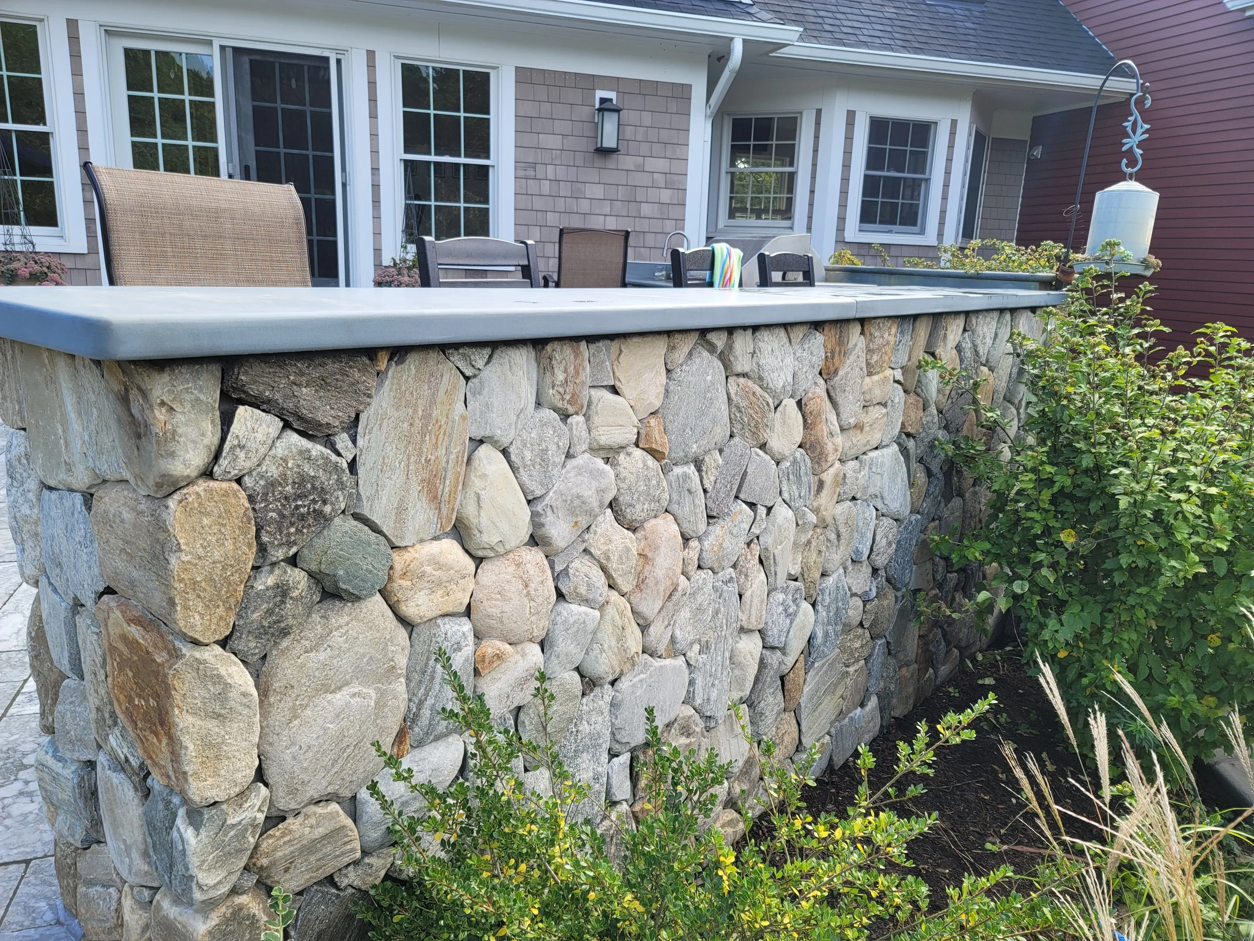 A backyard stone bar with a marble countertop, surrounded by patio furniture, plants, and a house with large windows and brick siding.