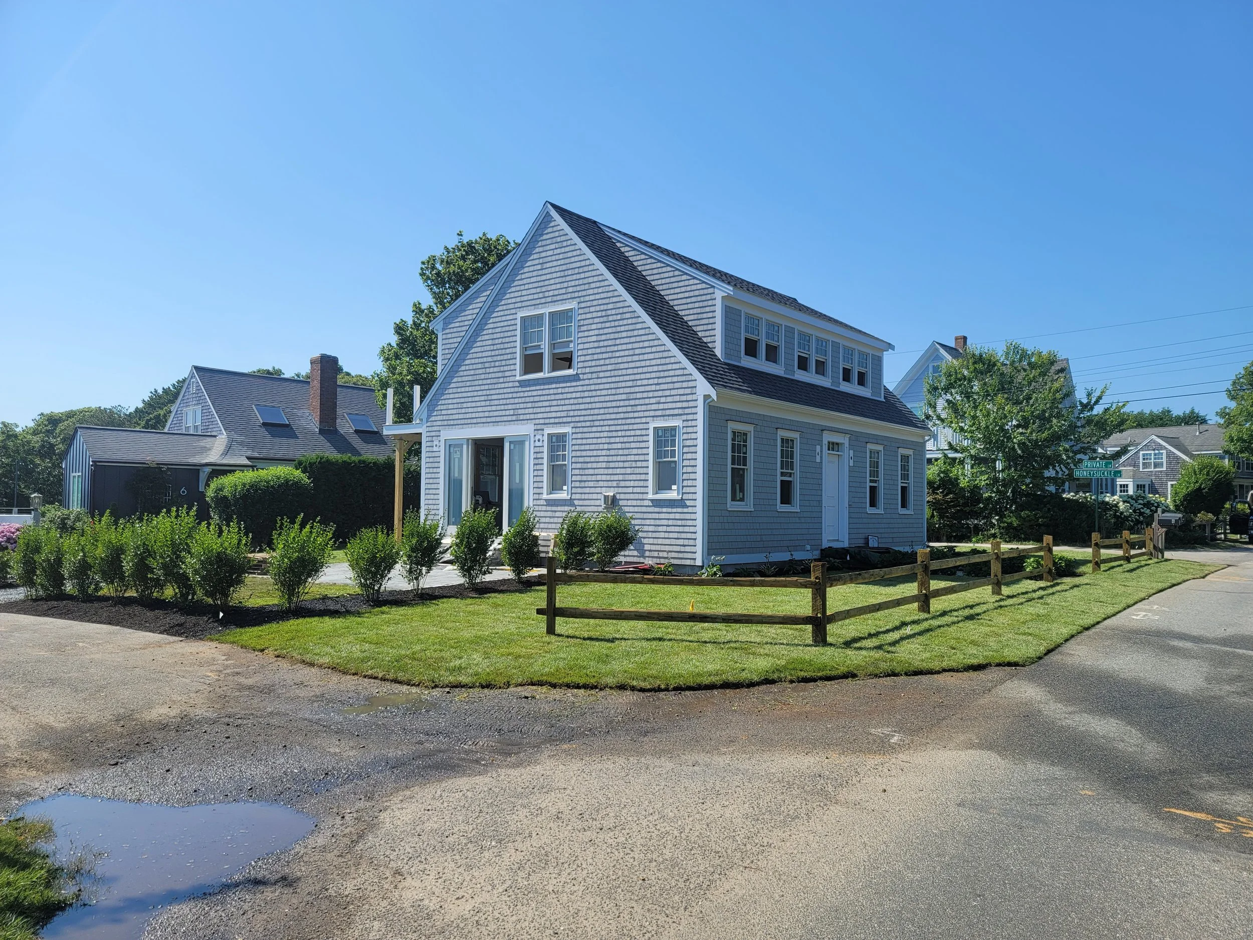A blue, multi-story house with a gabled roof and multiple windows, situated on a corner lot with a manicured lawn and small bushes, in a neighborhood with other houses and trees under a clear blue sky.