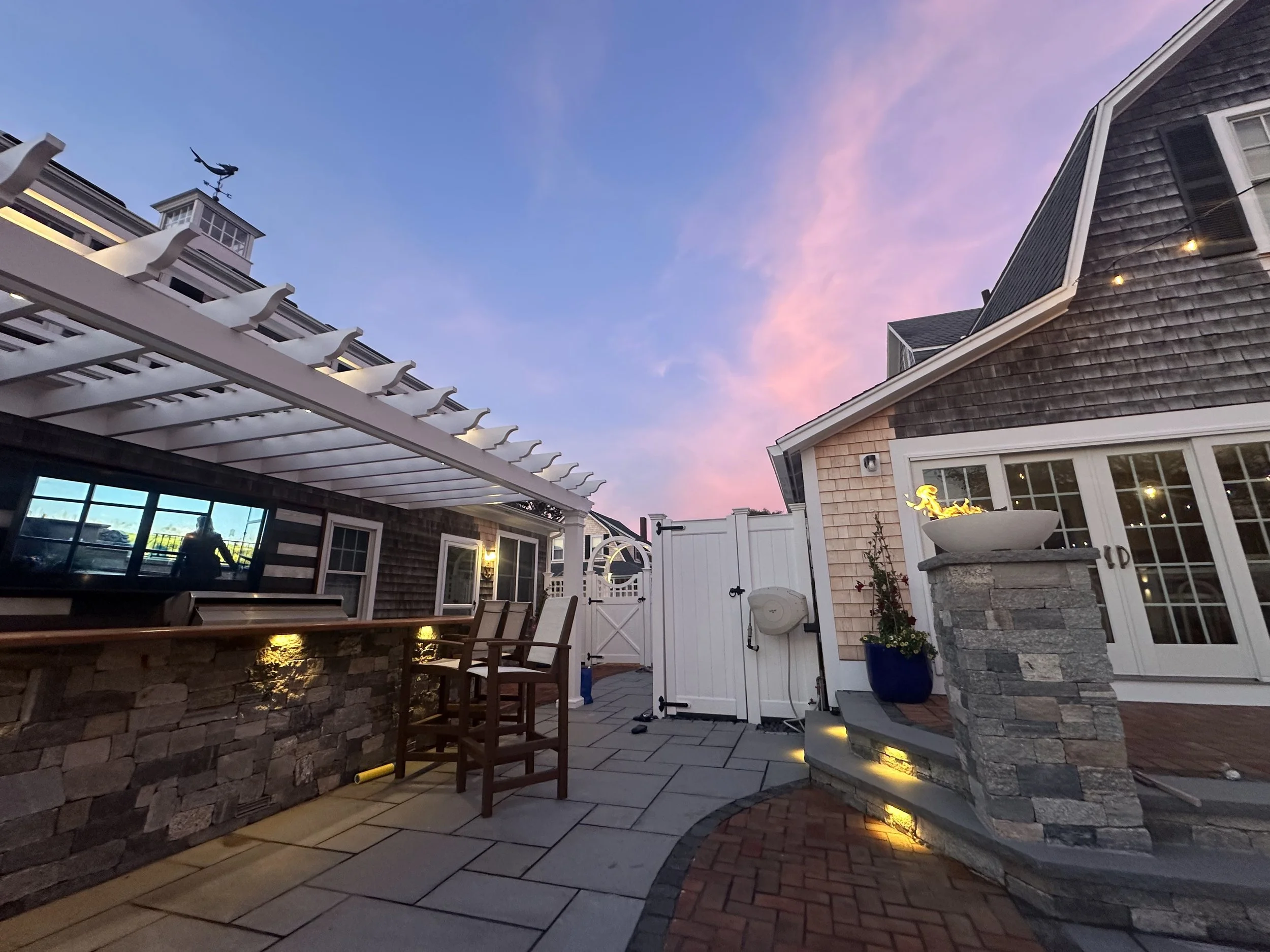 Backyard patio area with stone and brick features, outdoor seating, and a colorful sunset sky.