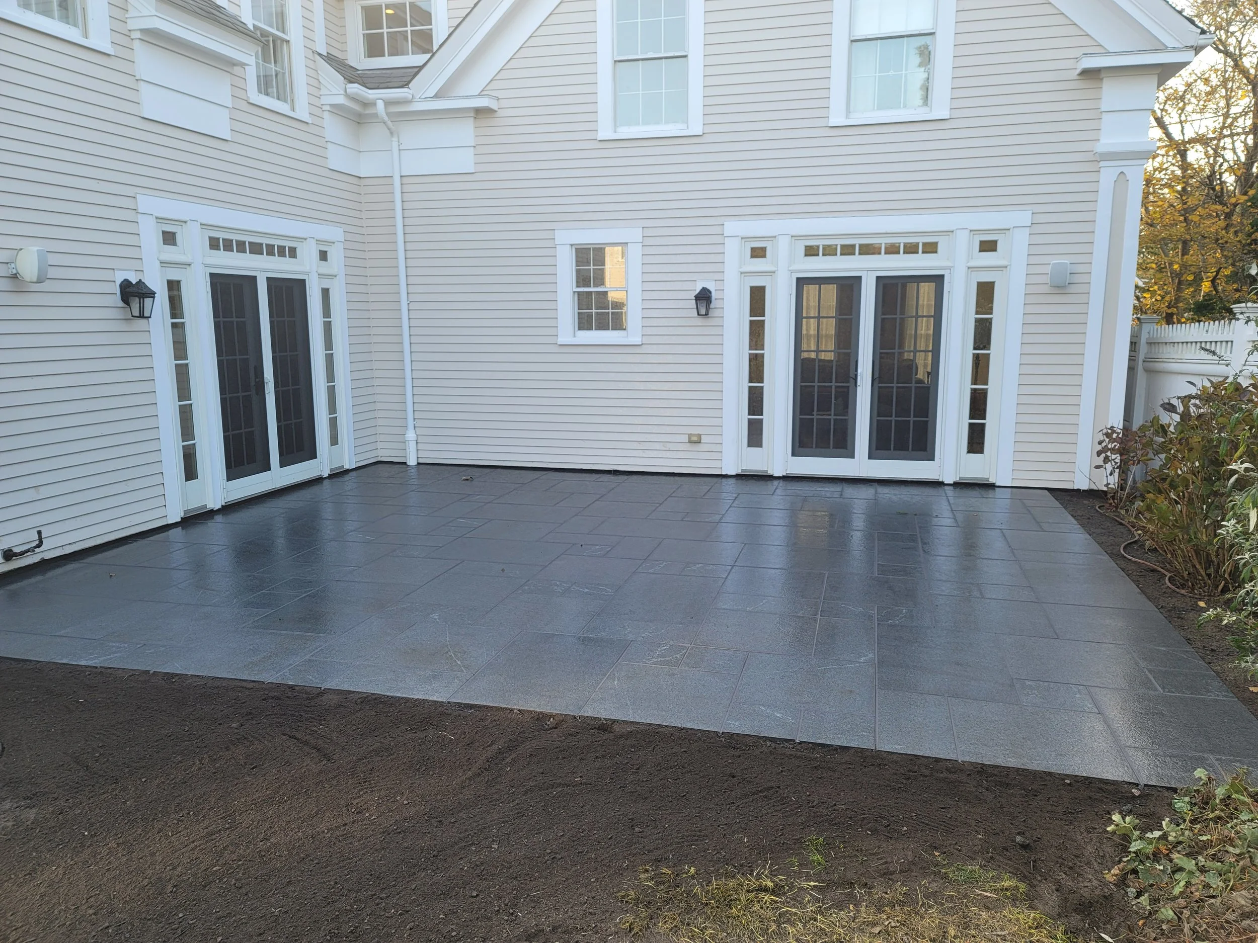 Backyard patio with dark gray tiles in front of a white house with siding and multiple windows and patio doors, surrounded by landscaping.