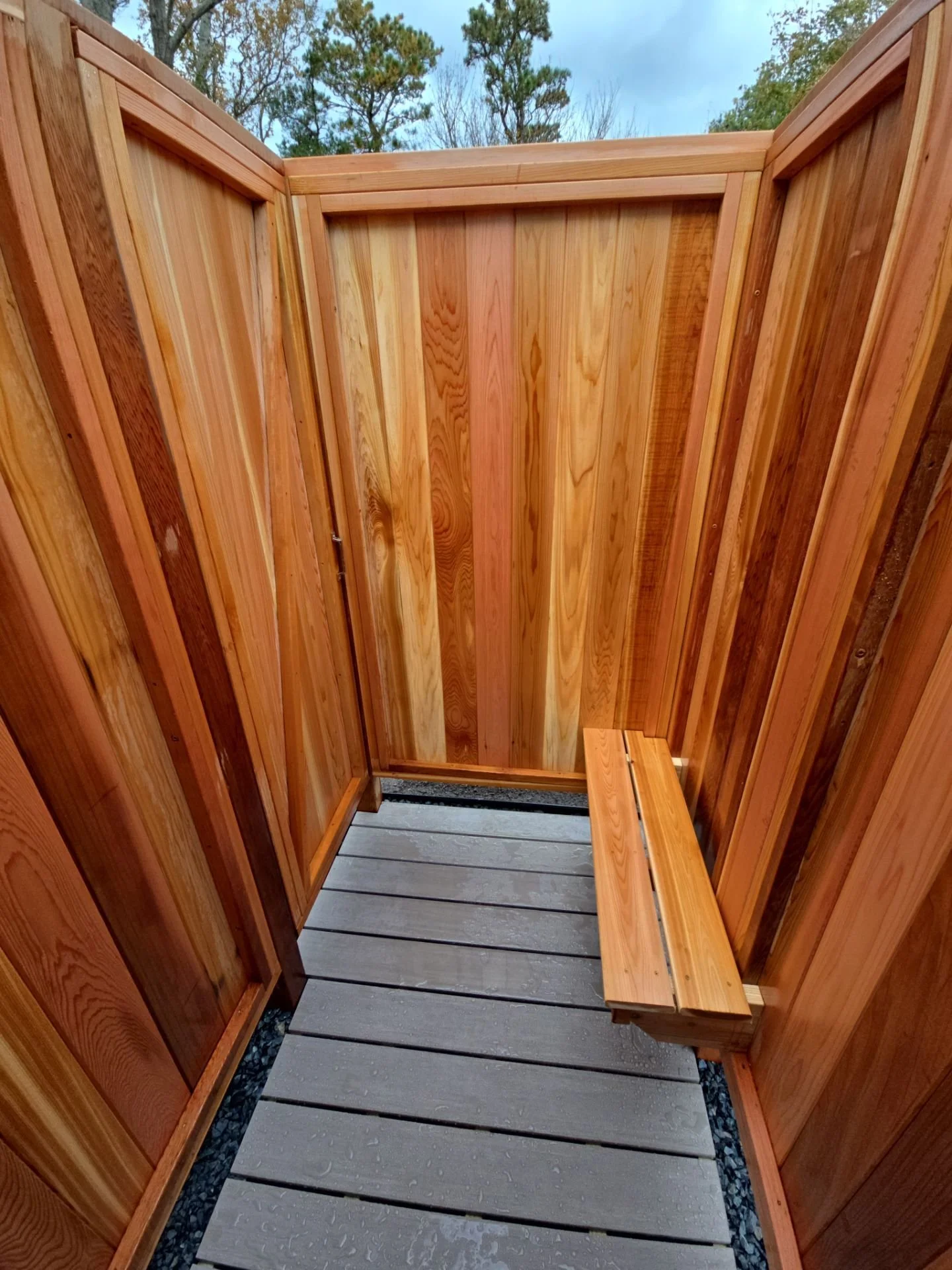 Small outdoor shower with wooden walls and a small wooden bench on a grey slatted floor, surrounded by trees.