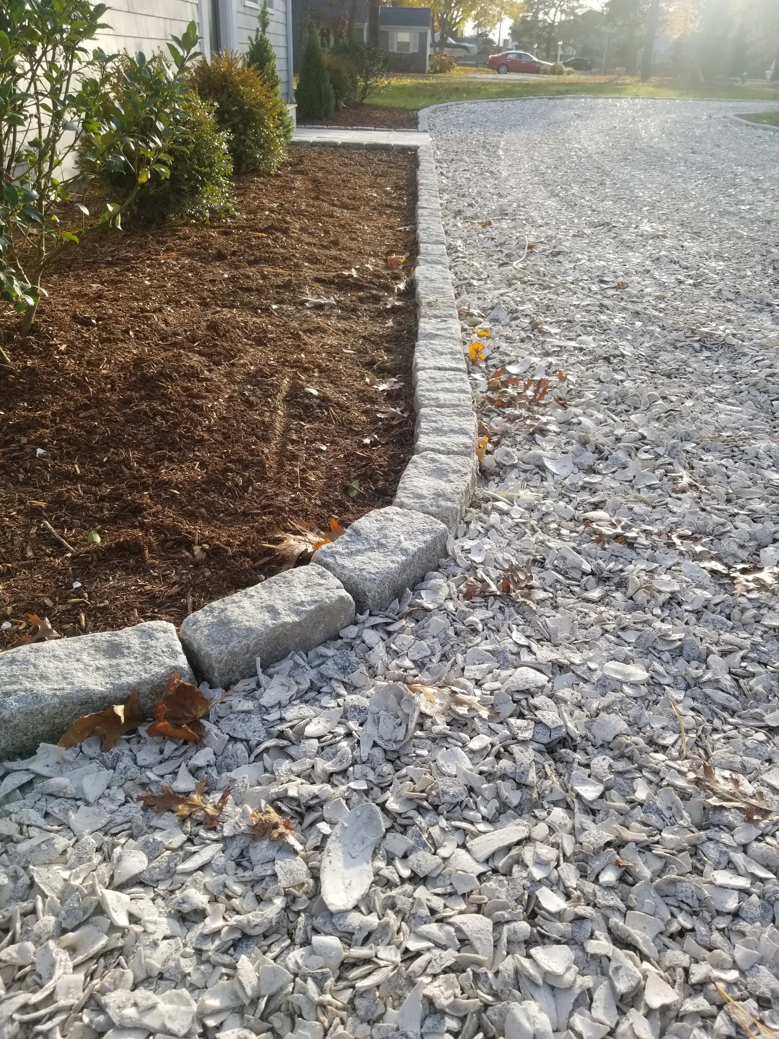 A gravel driveway with a curving border of large rocks, separating it from a garden bed with soil and bushes. Trees and houses are visible in the background on a sunny day.