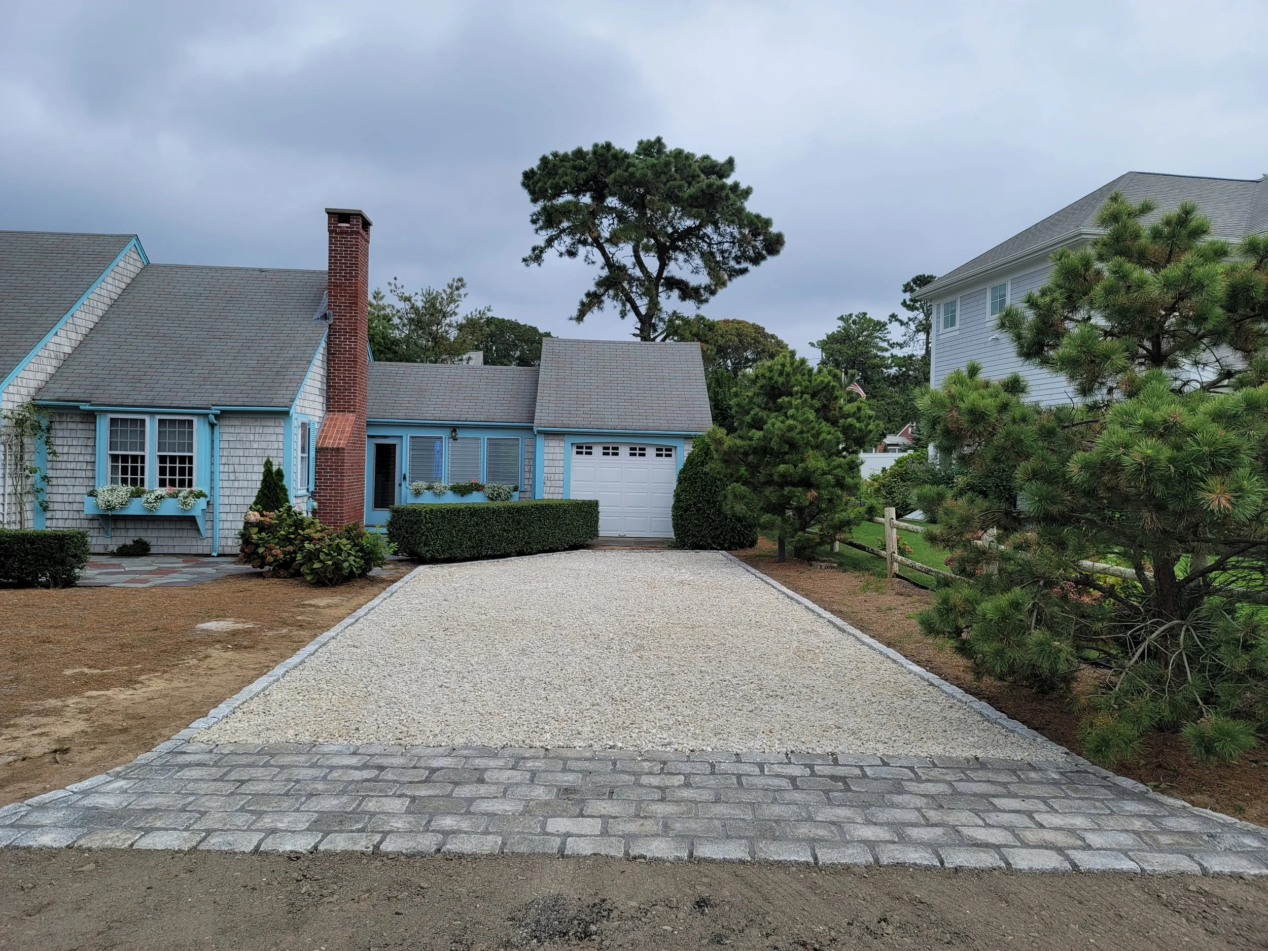 A driveway with a surface of gravel and cobblestone border leading to a house with a white garage door, blue siding, and a brick chimney. There are trees and shrubs on either side of the driveway.