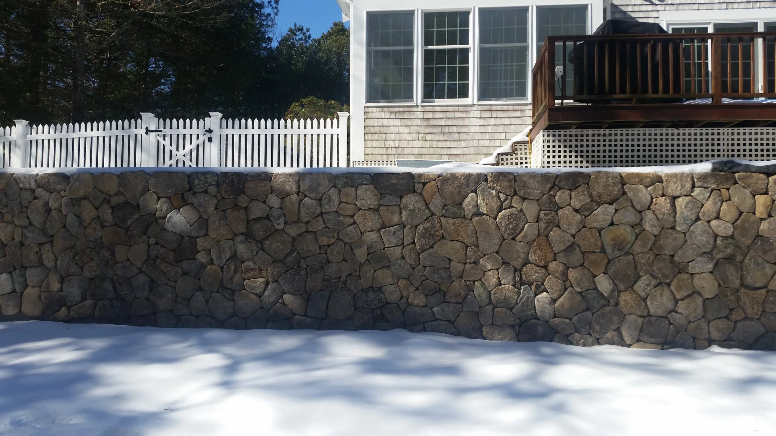 A backyard with a stone retaining wall, snow on the ground, a white fence, and a house with a wooden deck and large windows.