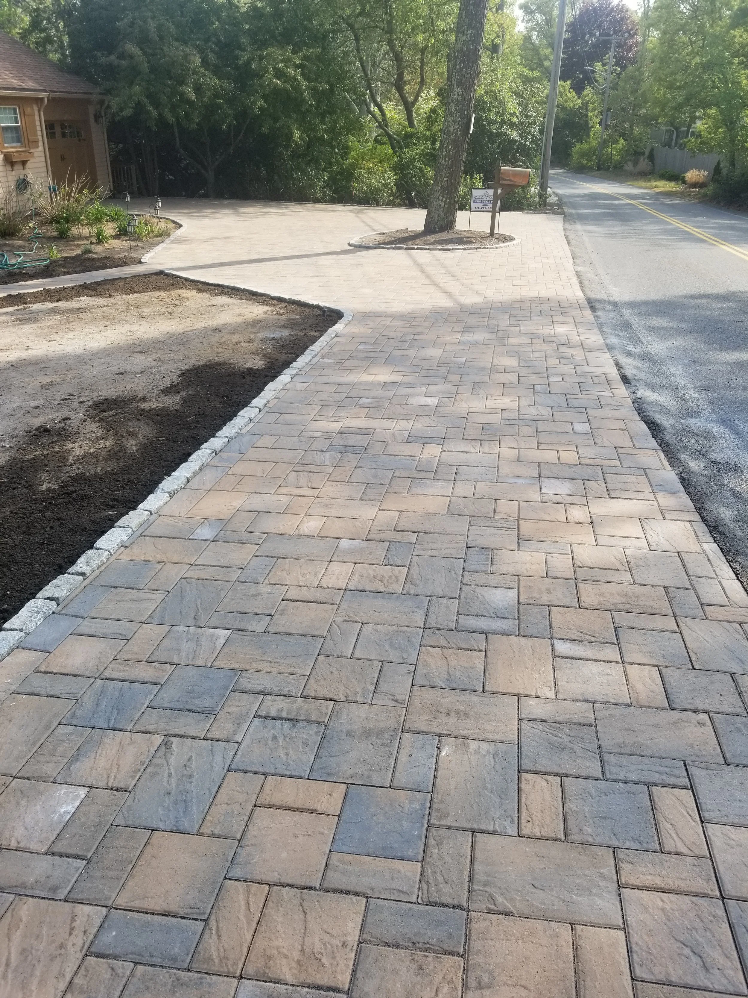 Newly paved brick sidewalk along a residential street, with a tree and mailbox at the corner, and a driveway to the left.