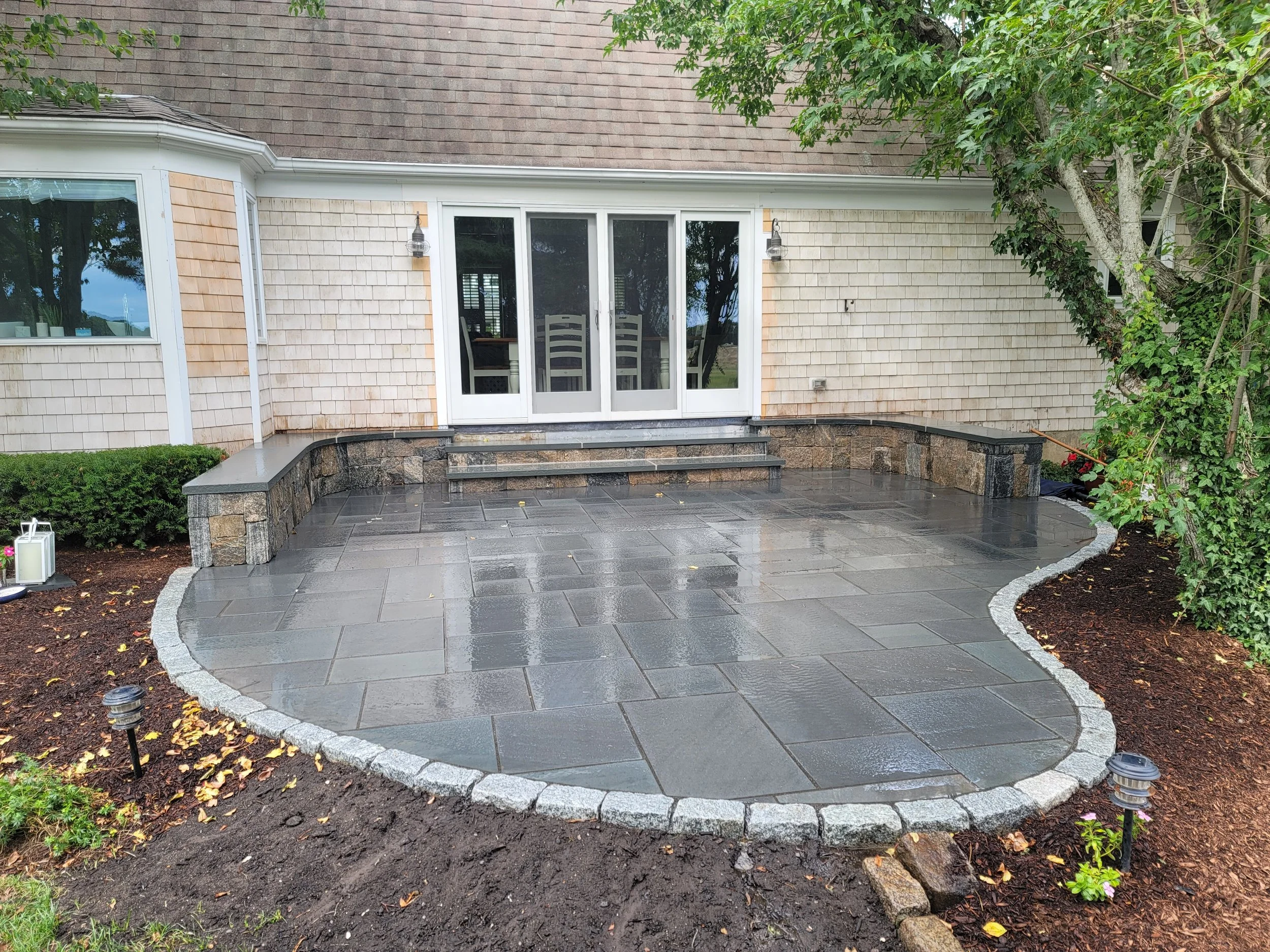 Backyard patio with wet stone tiles, steps leading up to a house with sliding glass doors, surrounded by greenery and landscaping lights.