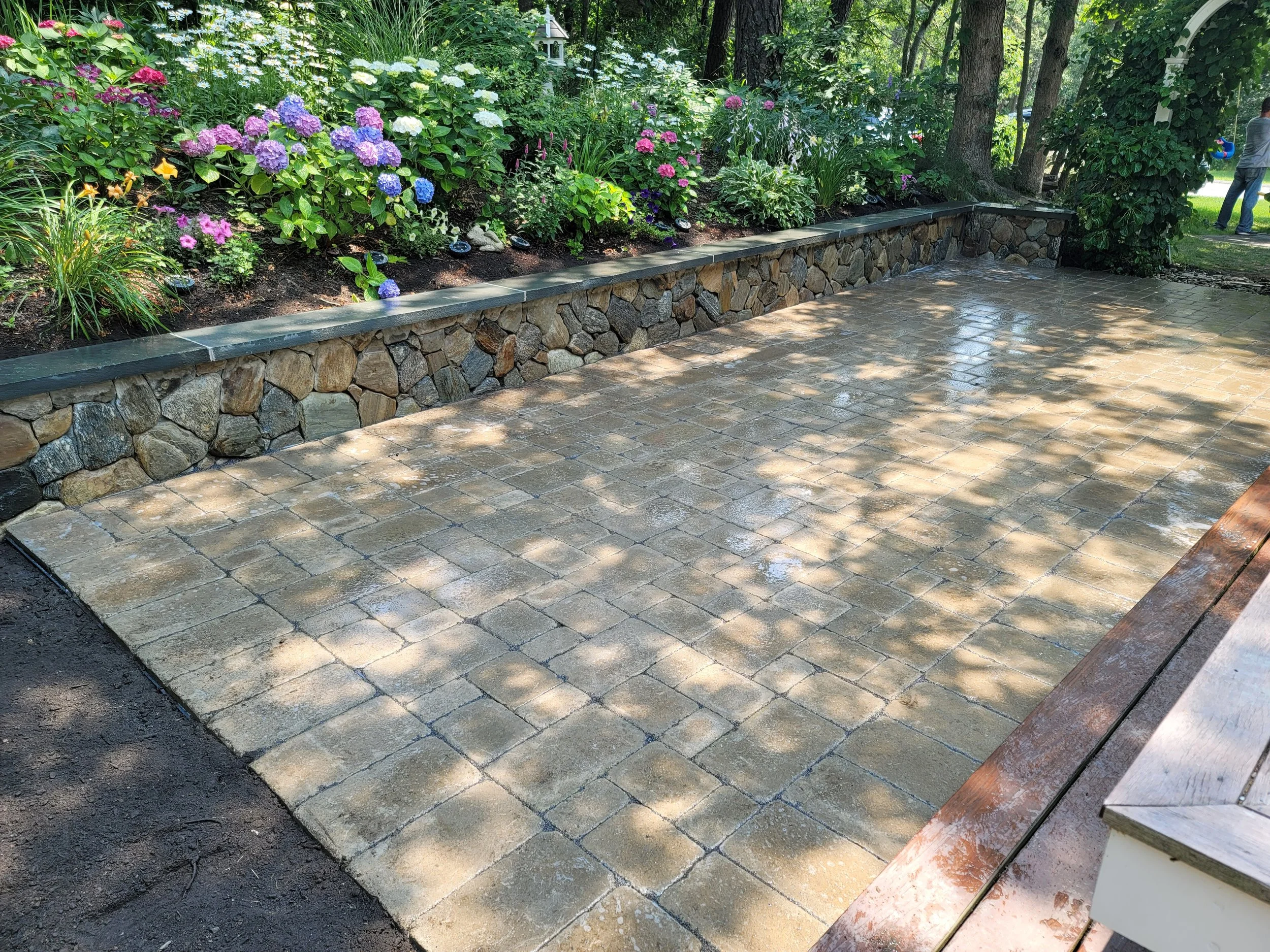 Stone-paved patio with a flower garden and trees in the background, and a person walking in the distance.