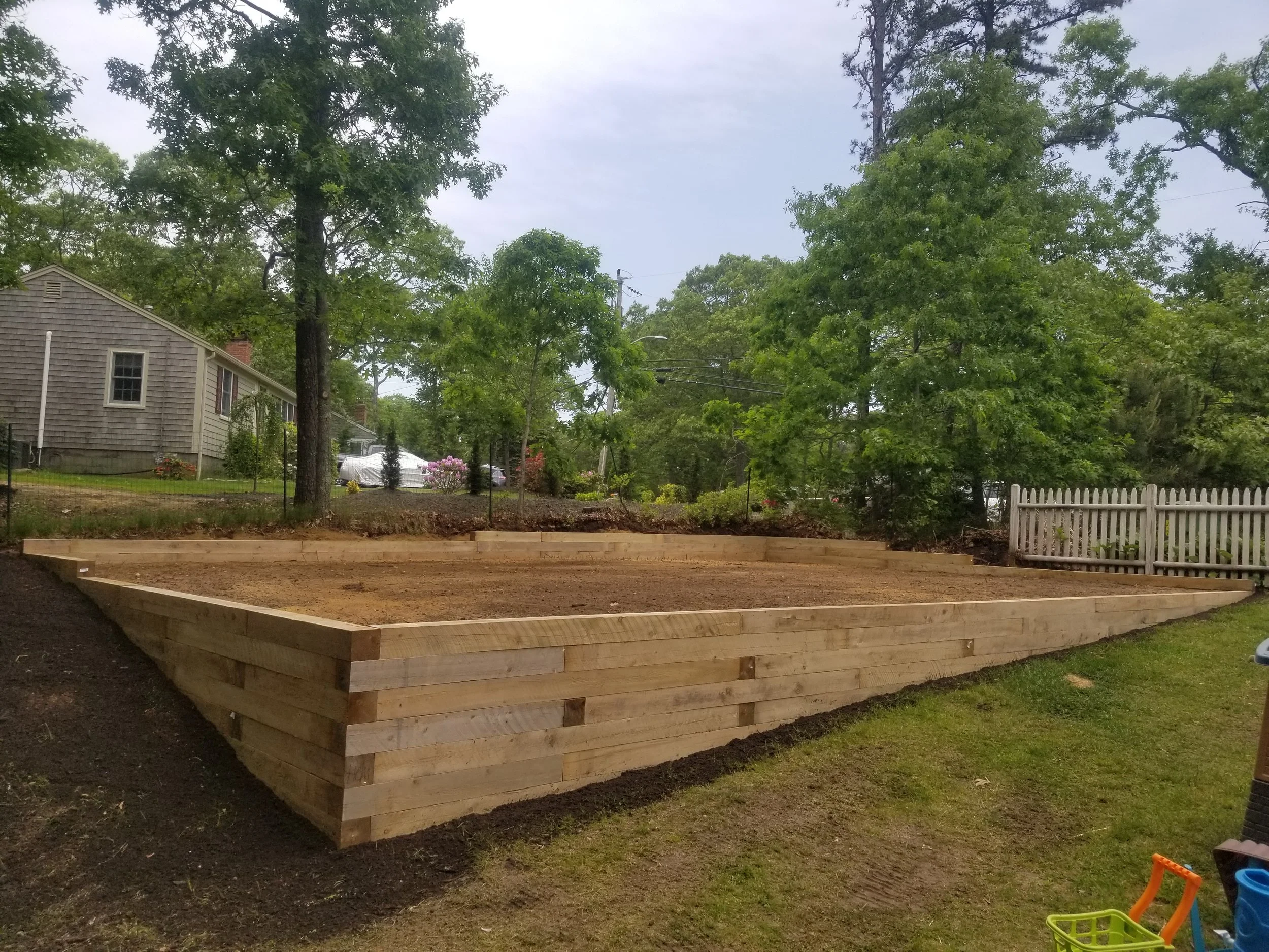 A backyard with a freshly built wooden retaining wall surrounding a level patch of dirt, green grass, trees, and a white picket fence in a suburban neighborhood.