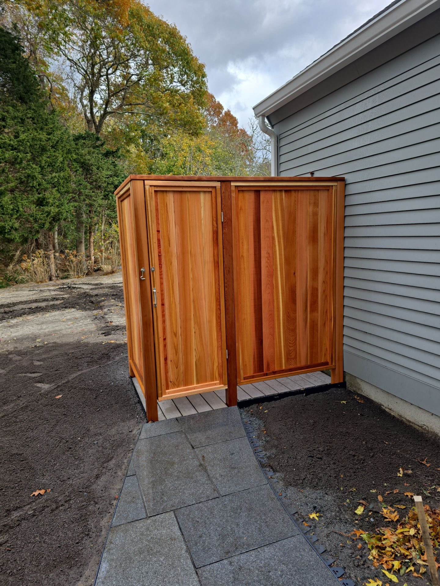 Wooden outdoor shower enclosure next to house, with gray siding, paving stones in front, trees in the background, and cloudy sky.
