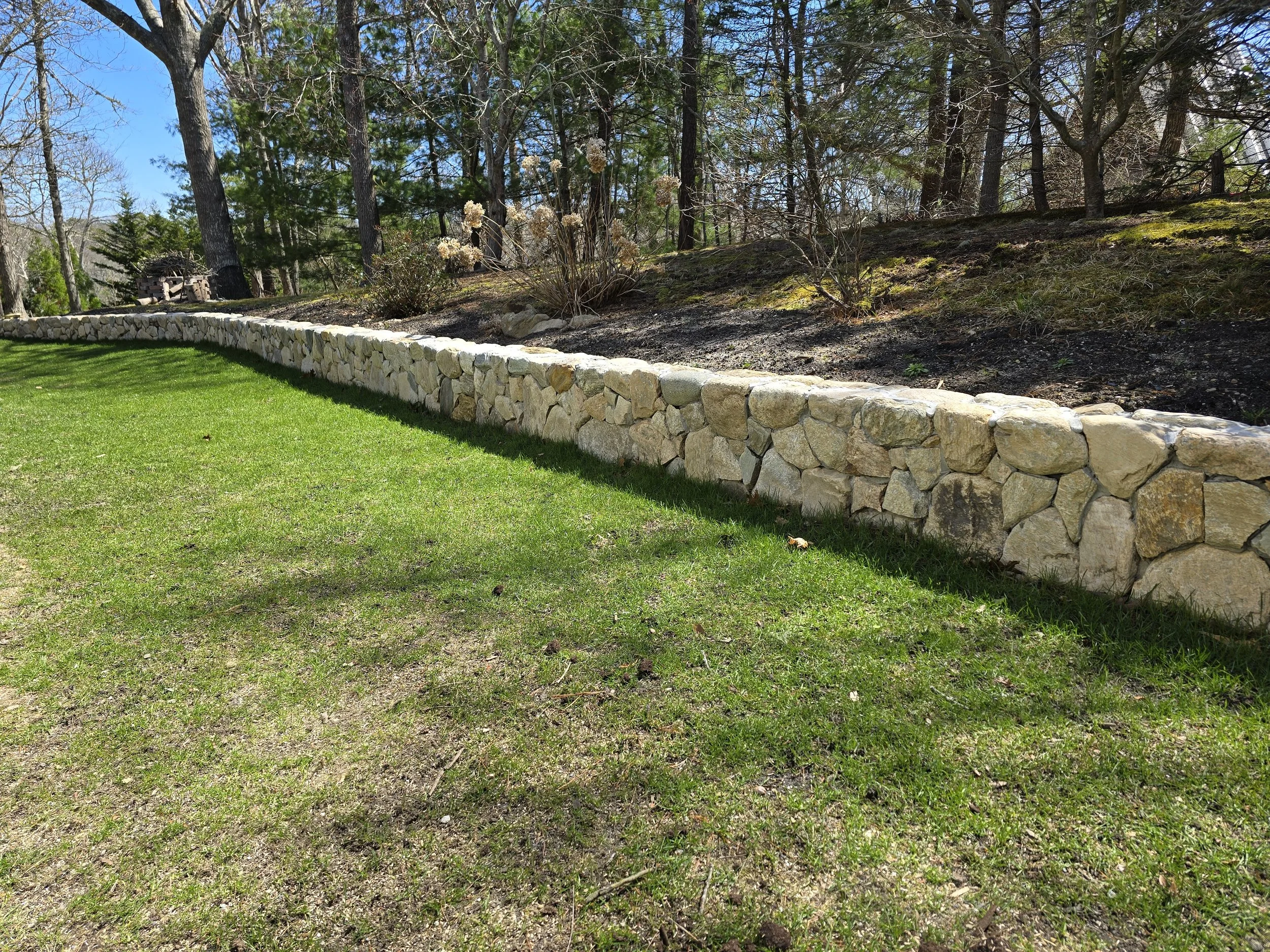 A stone retaining wall separates a well-maintained grassy lawn from a wooded area with trees and bushes.