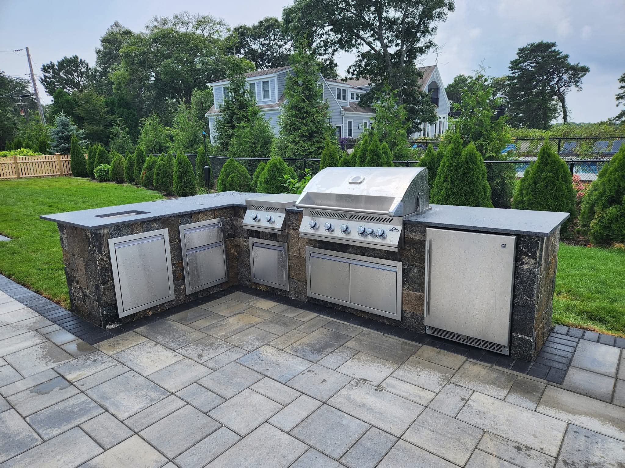Outdoor kitchen with built-in grill, sink, and storage cabinets, surrounded by a landscaped backyard with a grassy lawn, manicured bushes, and trees.