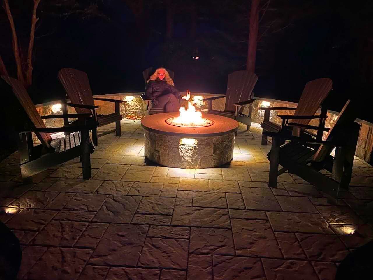 A woman sitting around a fire pit on a patio at night, with five wooden chairs positioned around it. The scene is illuminated by the fire and small ground lights, with trees and darkness in the background.