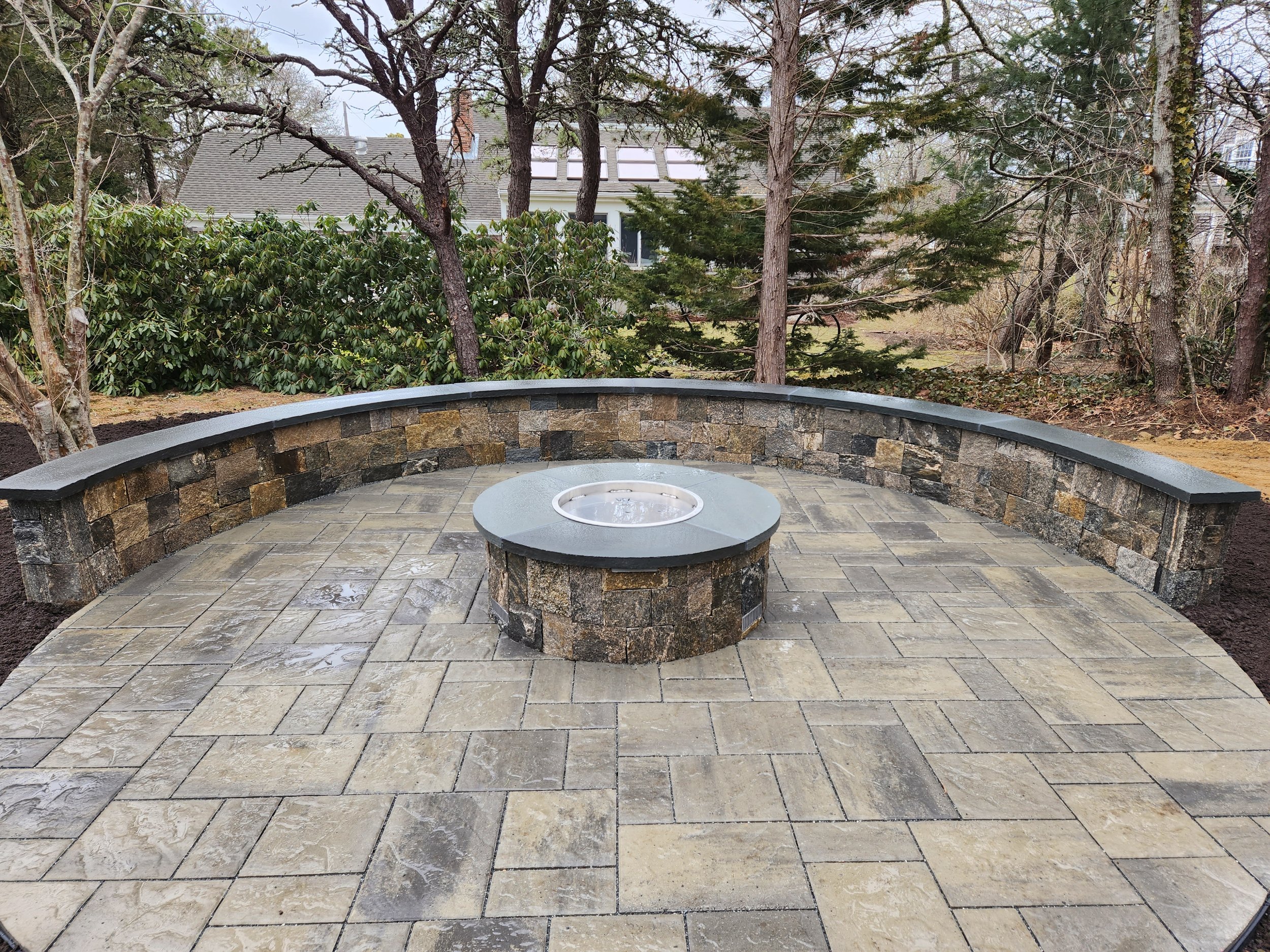A circular outdoor patio area with a stone fire pit in the center and a curved stone wall around the perimeter. The patio is paved with rectangular stone tiles, and there are trees and houses in the background.