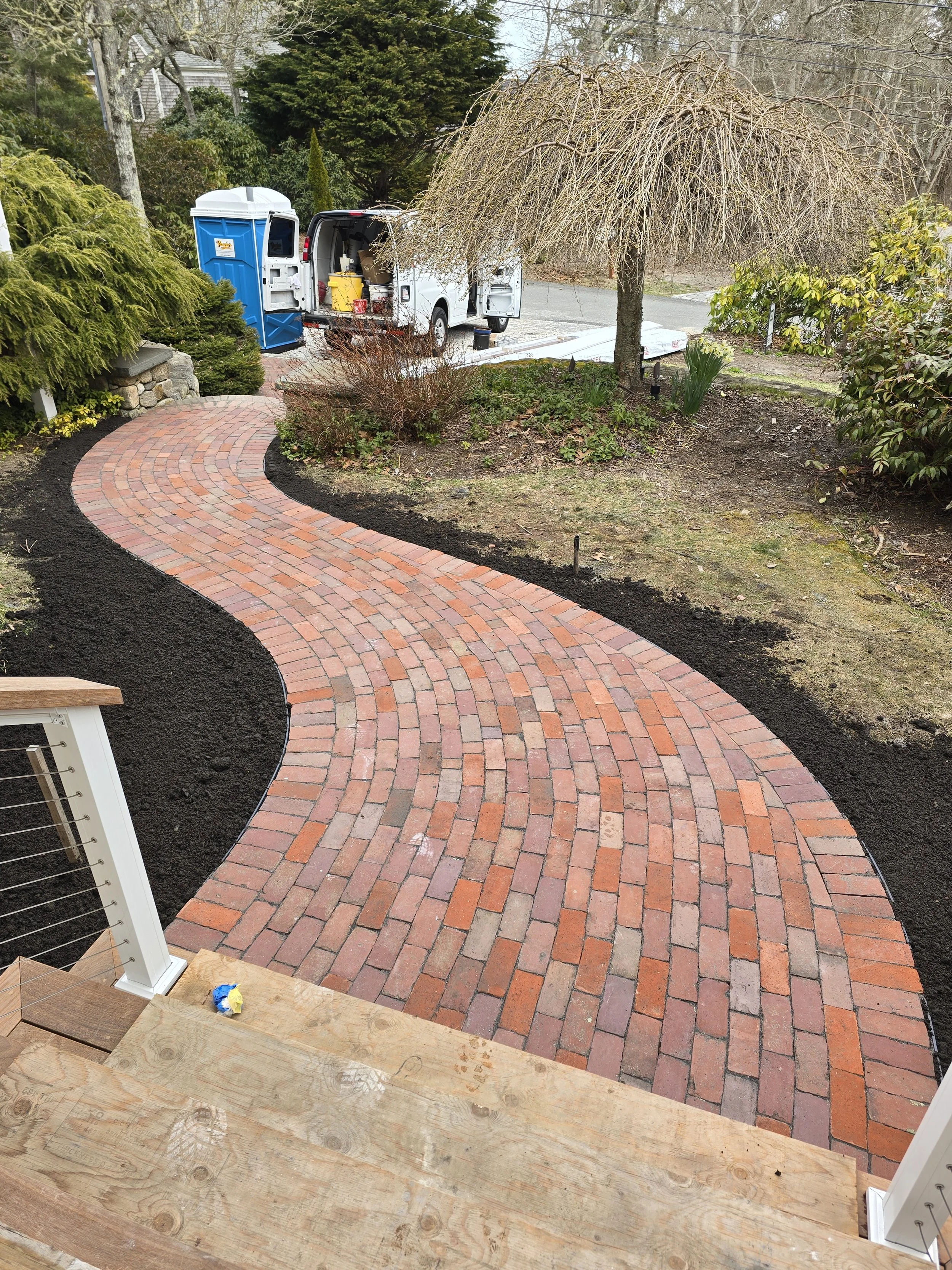 Newly constructed brick pathway with a curved design leads from a wooden porch to a driveway with a white van, surrounded by a garden with various plants and a leafless tree.