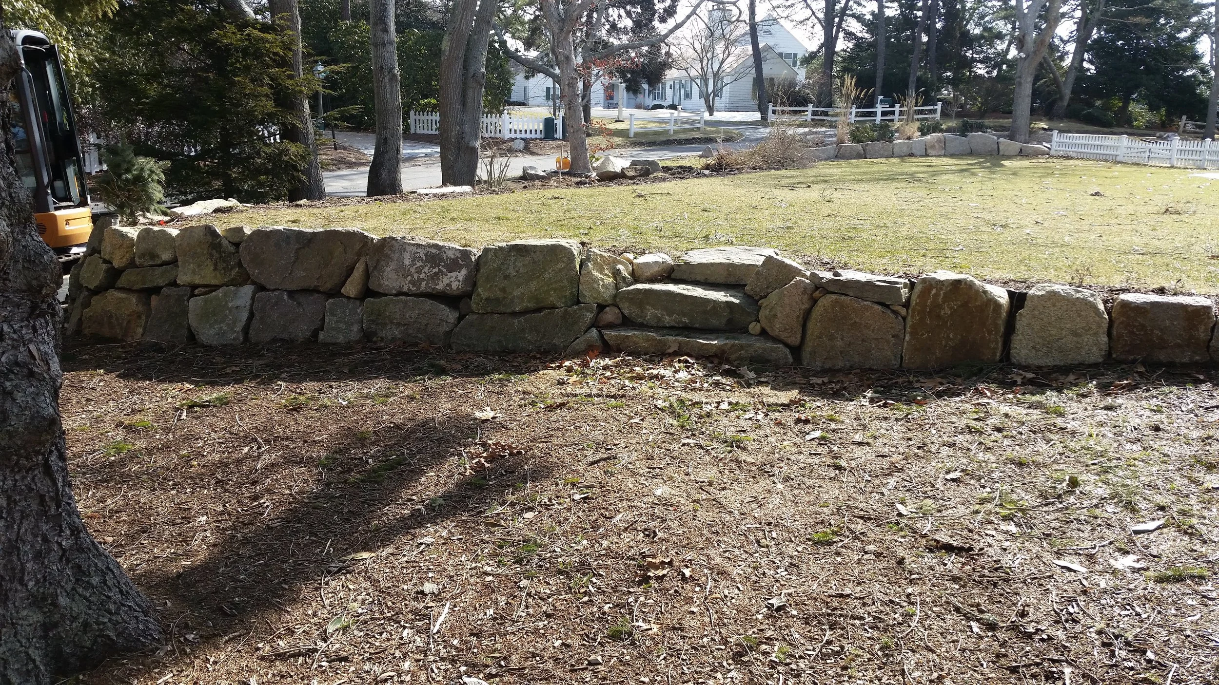 A stone retaining wall in a yard with trees, grass, and houses in the background.