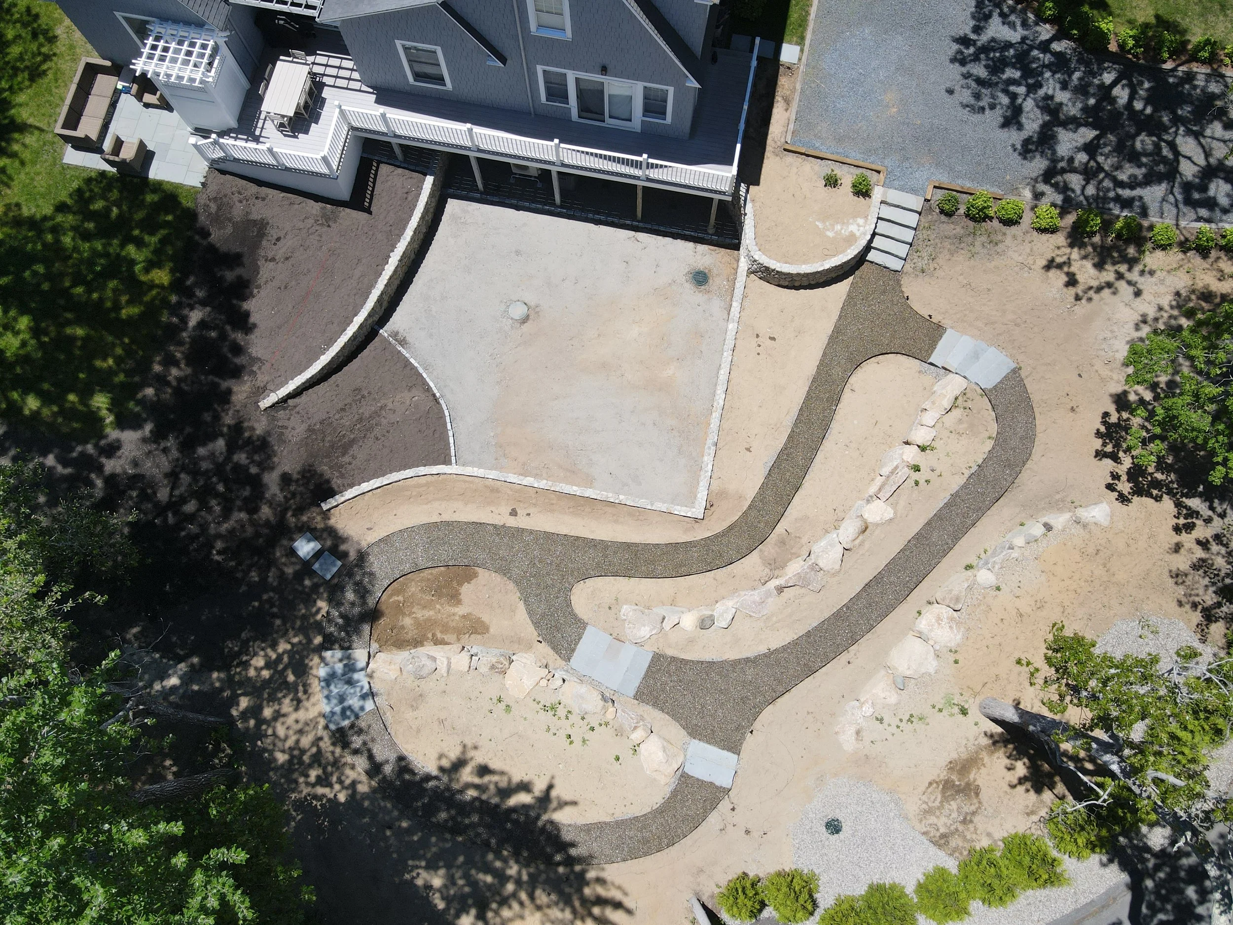 An aerial view of a backyard with a winding pathway, partially paved patio, and a house with a deck. The yard has trees, bushes, and areas of bare soil and gravel.