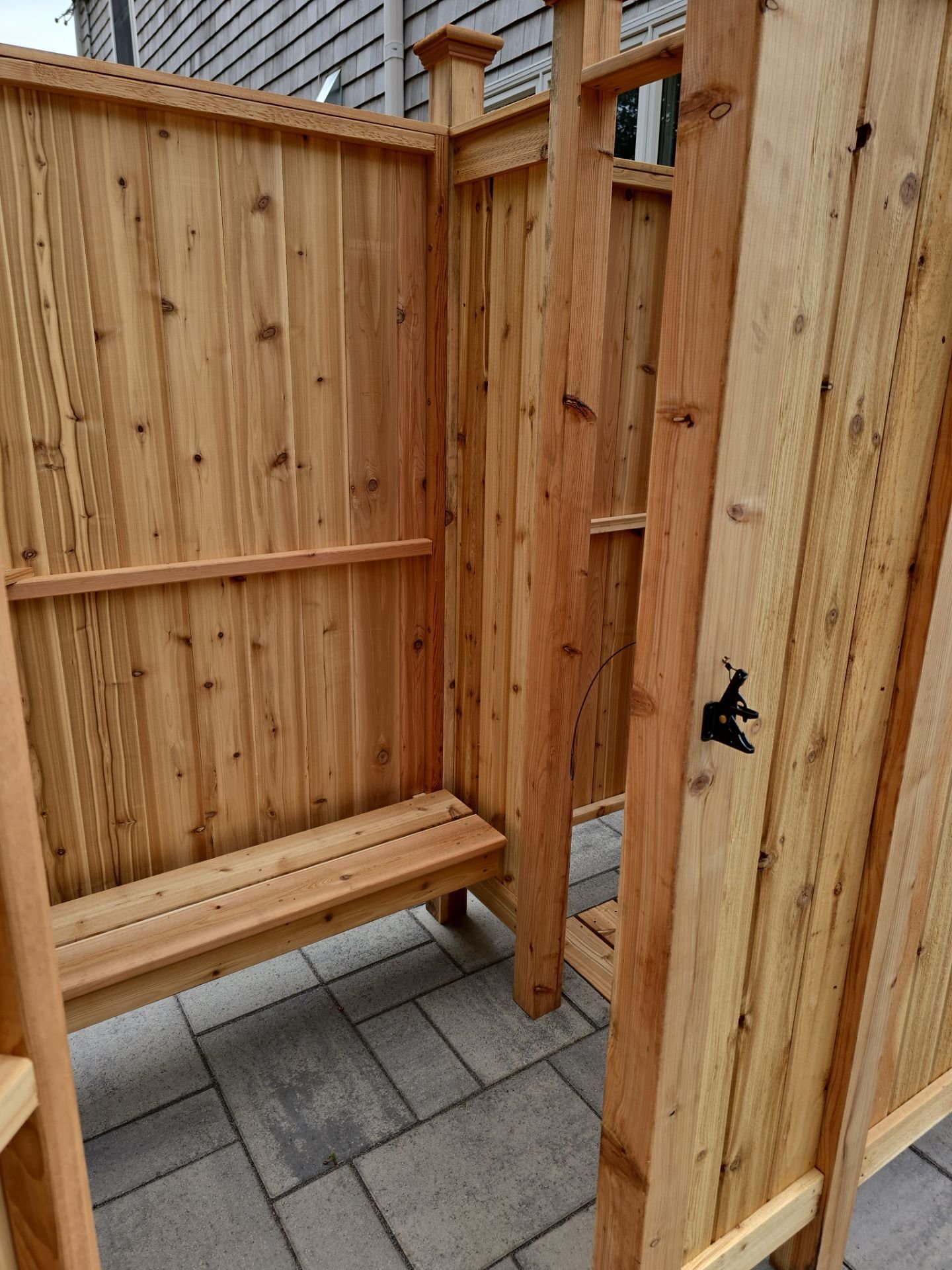 Unfinished wooden outdoor shower with a bench, installed on a paved surface, in front of a house with gray siding.
