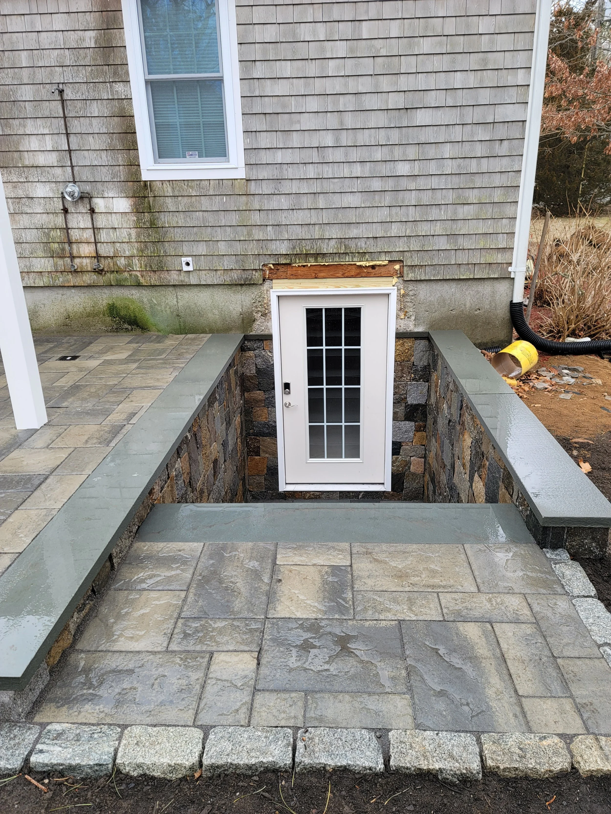 Backyard patio with stone paving and new framed door leading to basement, with an outside wall of a house, window, and some construction materials and plants nearby.