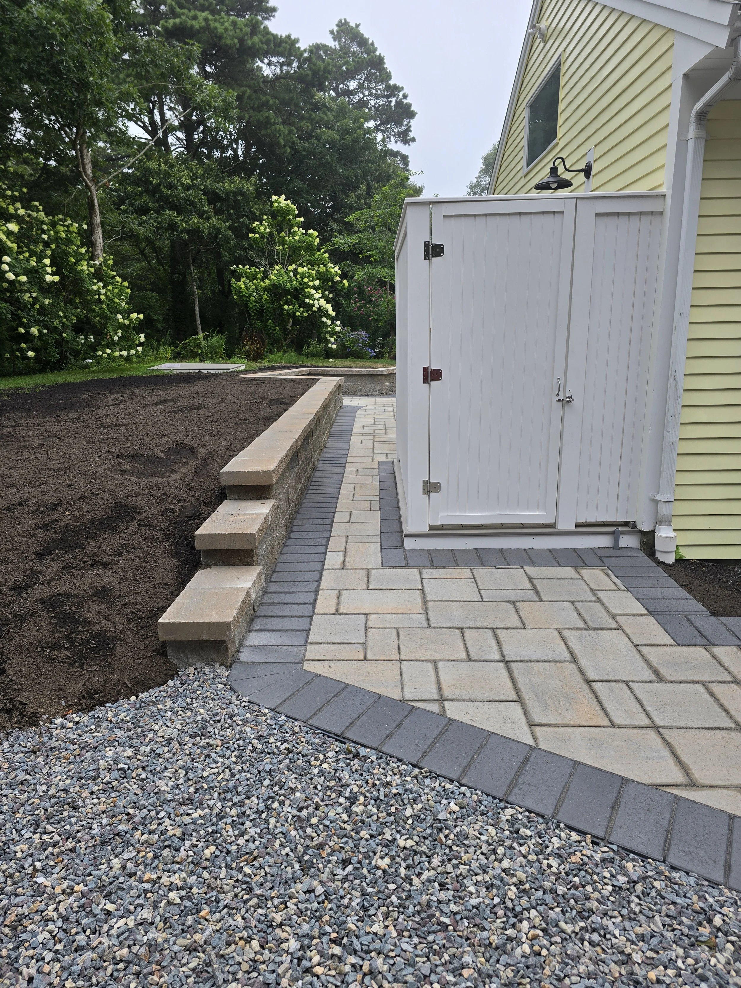 A backyard patio area with a paved walkway, a step-up brick wall, a shed with a door, and surrounding trees and shrubs.
