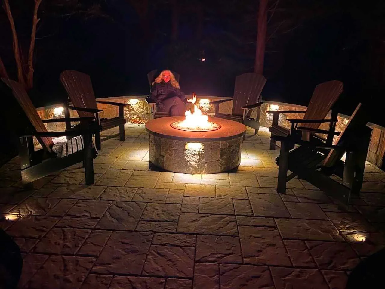 Person sitting on a chair around a fire pit at night, with five empty chairs arranged in a semi-circle.