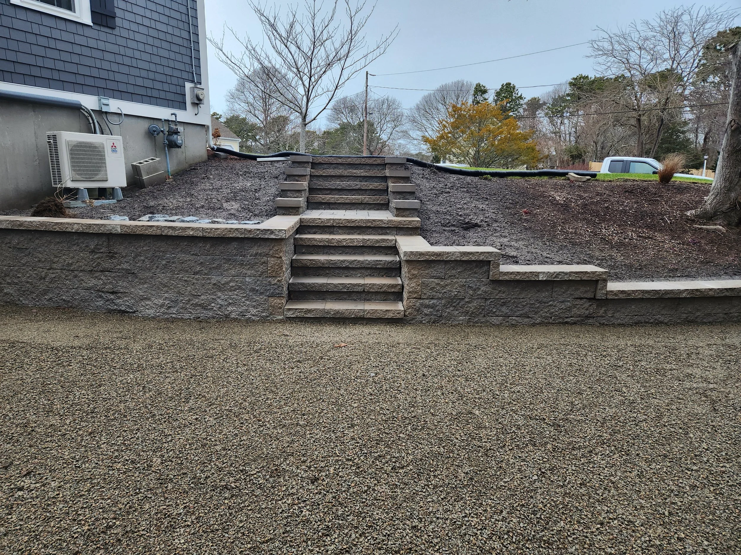 A set of outdoor concrete steps with a stone retaining wall, leading from the yard to the house elevation. The yard area to the right is unfinished soil, and there are trees and a parked vehicle in the background.