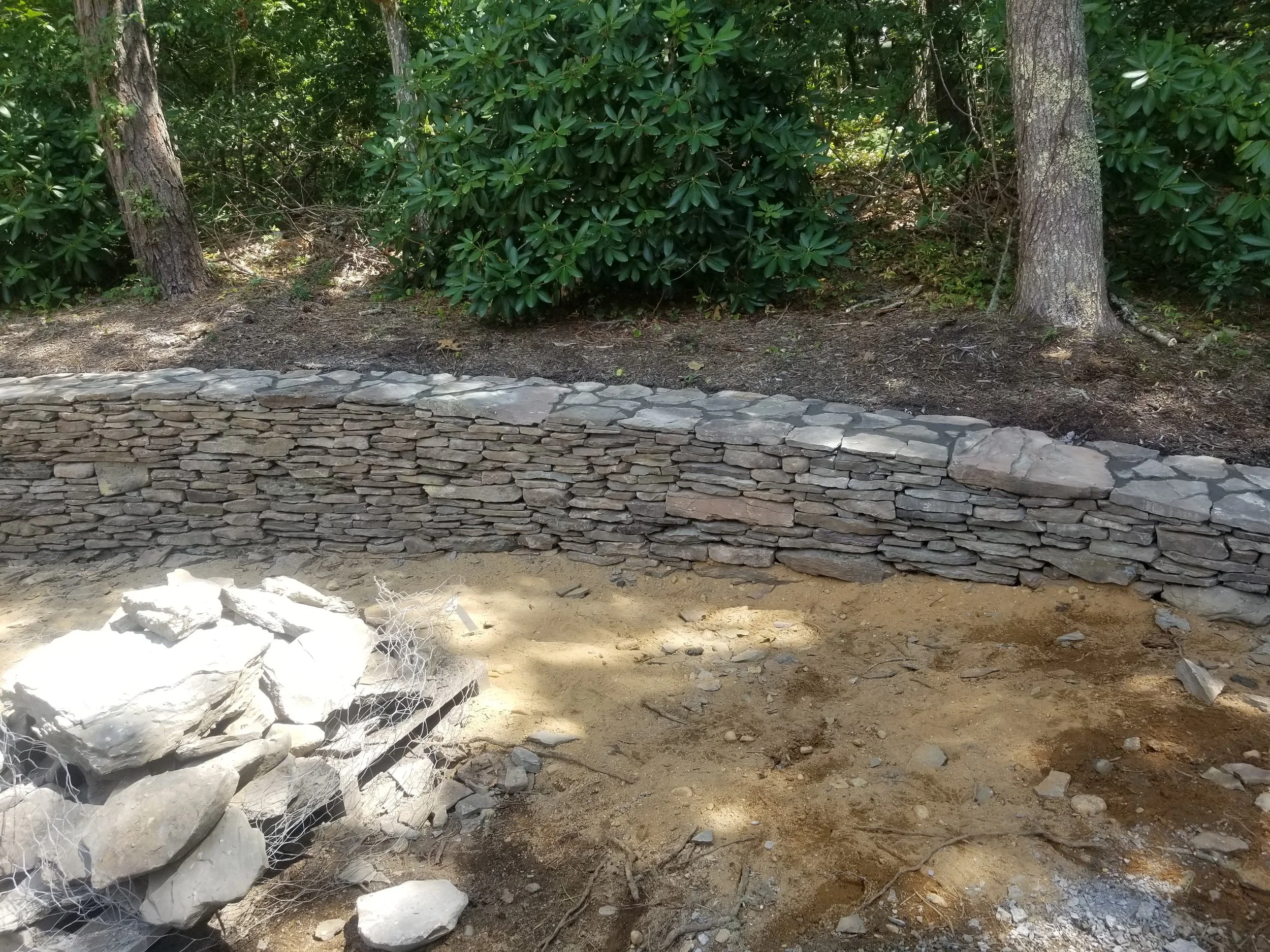 A stone retaining wall in a wooded outdoor area with trees and bushes in the background.