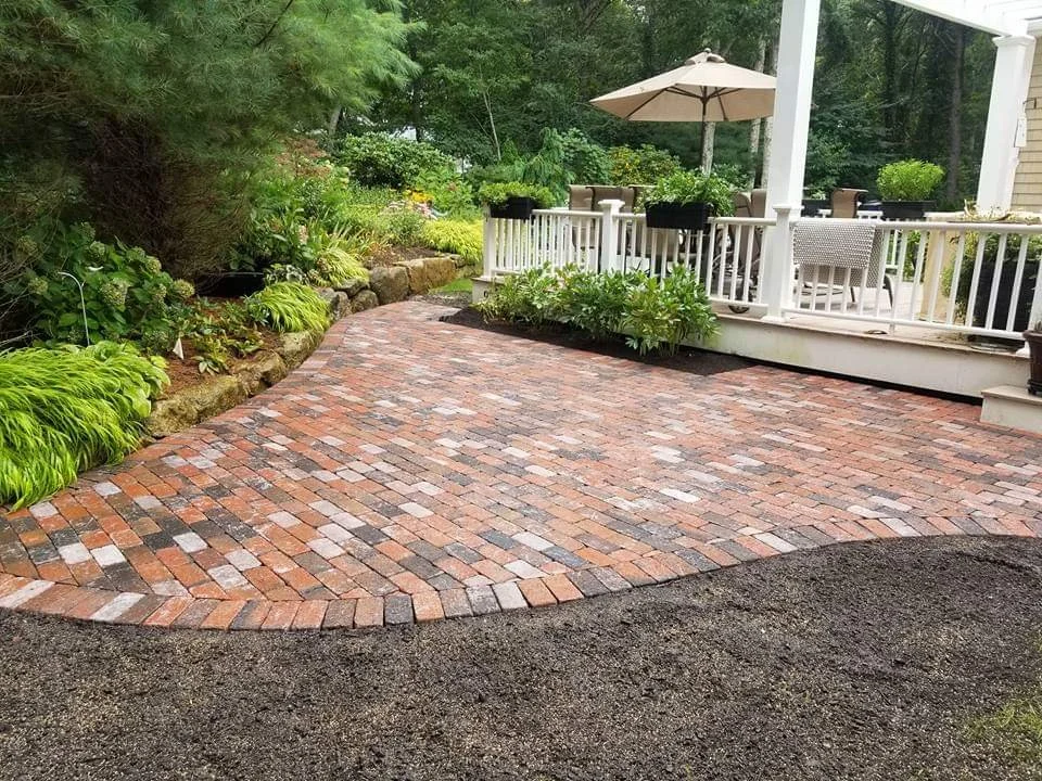 A backyard patio made of red and gray bricks, bordered by lush green plants and trees, with a white porch and patio furniture including an umbrella.