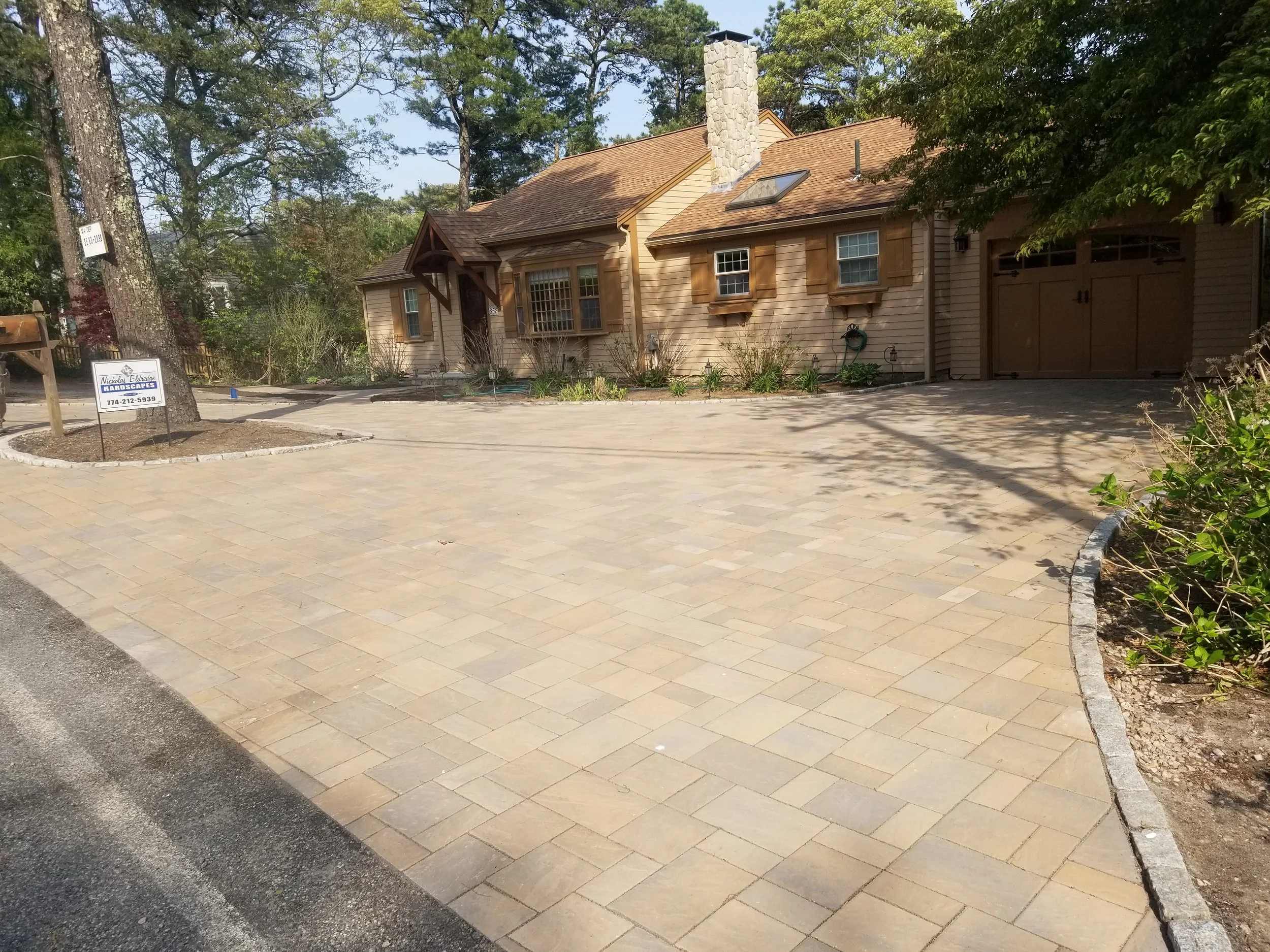 A house with a paved driveway and garden area, surrounded by trees, with a sign in the front yard for a landscaping business.