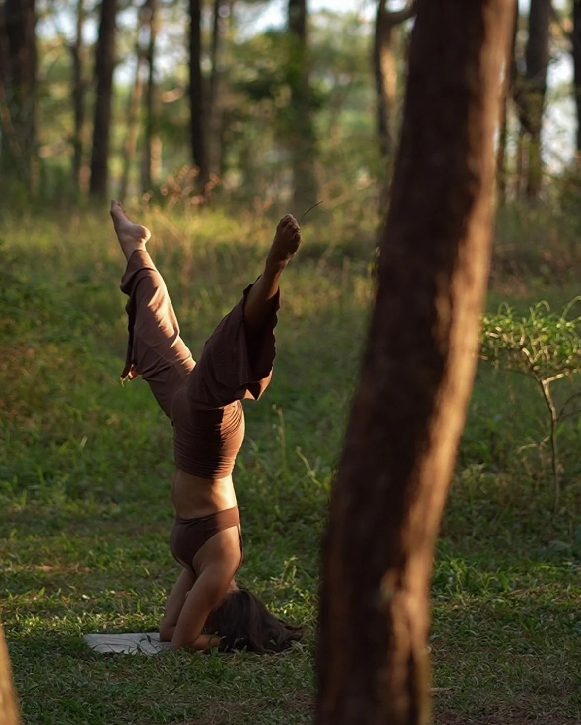 A woman practicing yoga in a forest during sunset, performing a headstand with legs extended upward.
