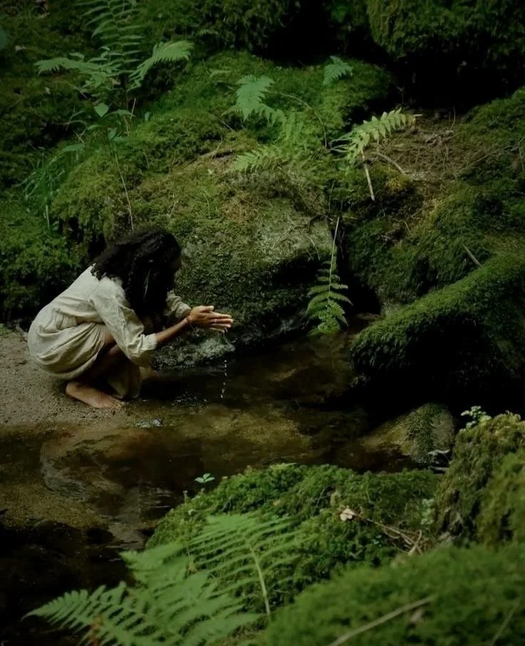 A woman kneeling by a small stream in a lush green forest, with moss-covered rocks and ferns surrounding her.